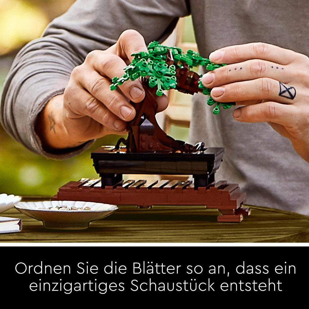 Hände, die einen Lego-Bonsai-Baum zusammensetzen, grüne Blätter auf dem braunen Stamm, auf Holzsockel.