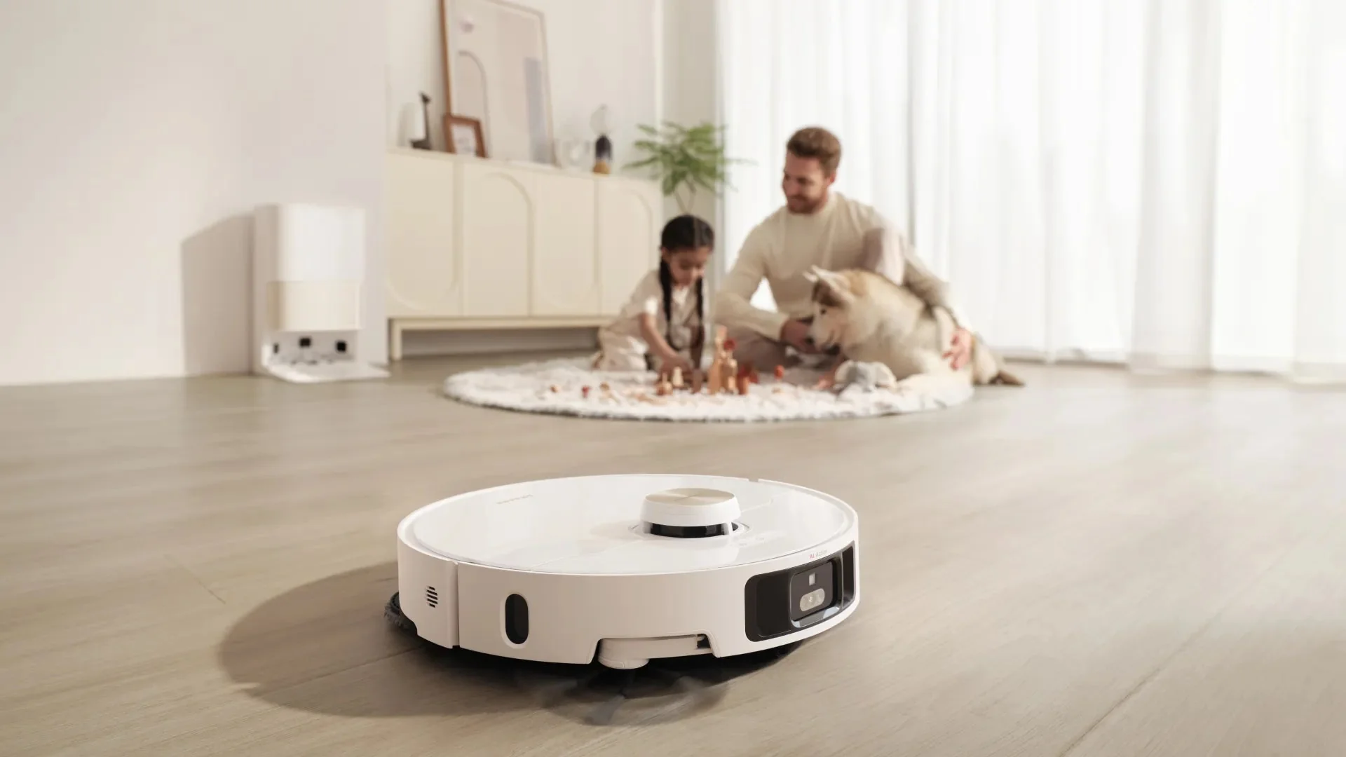 Fehér robotporszívó tisztít egy nappalit. Egy család játszik a közelben egy kutyával.
