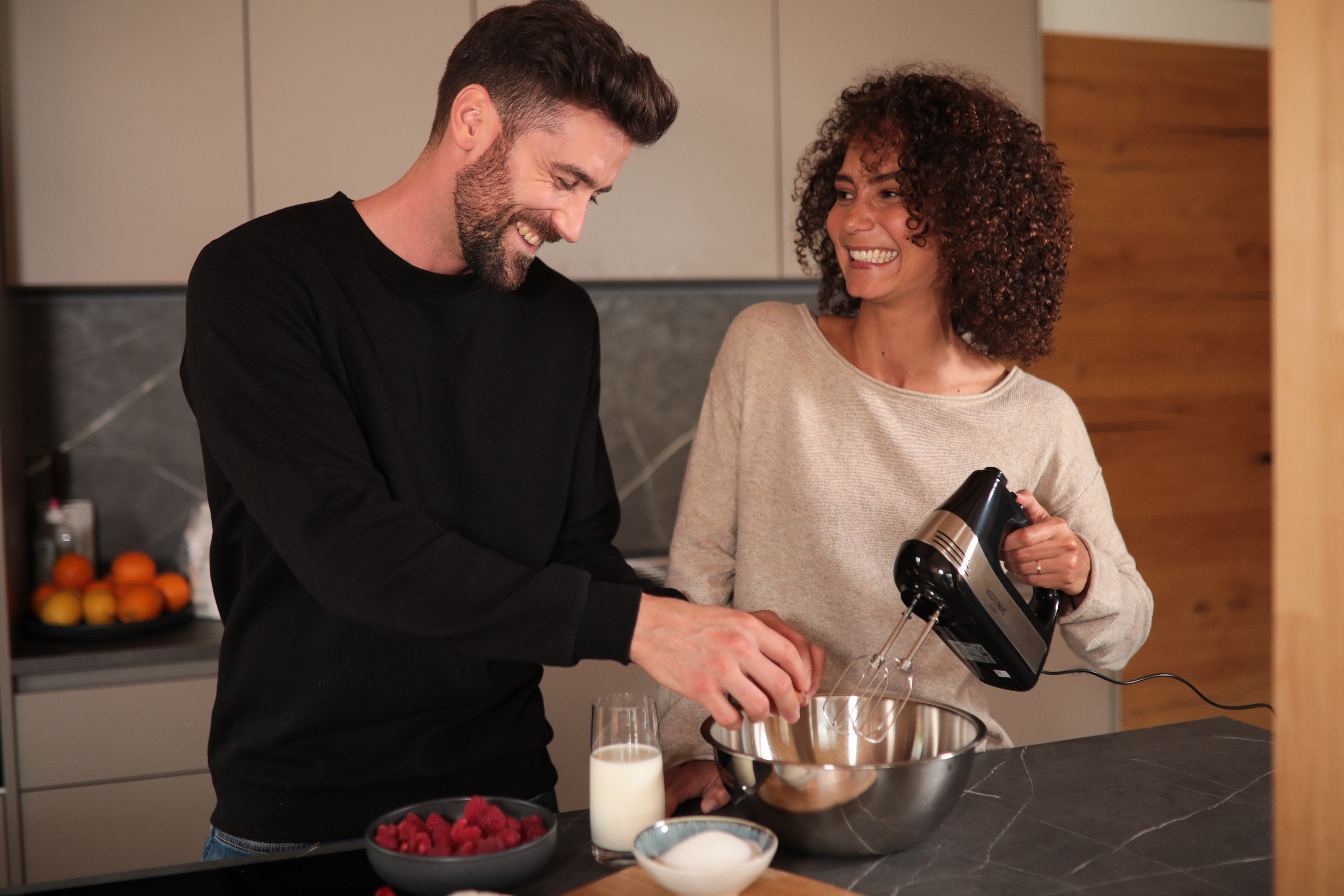 Una pareja en la cocina, hombre rompiendo un huevo en un bol, mujer usando una batidora.