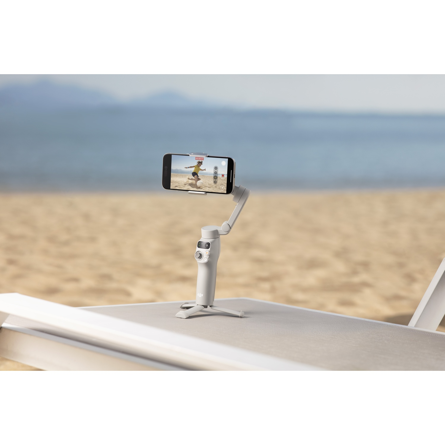 Un gimbal bianco che tiene un telefono, su una sedia. Una spiaggia e acqua blu sullo sfondo.