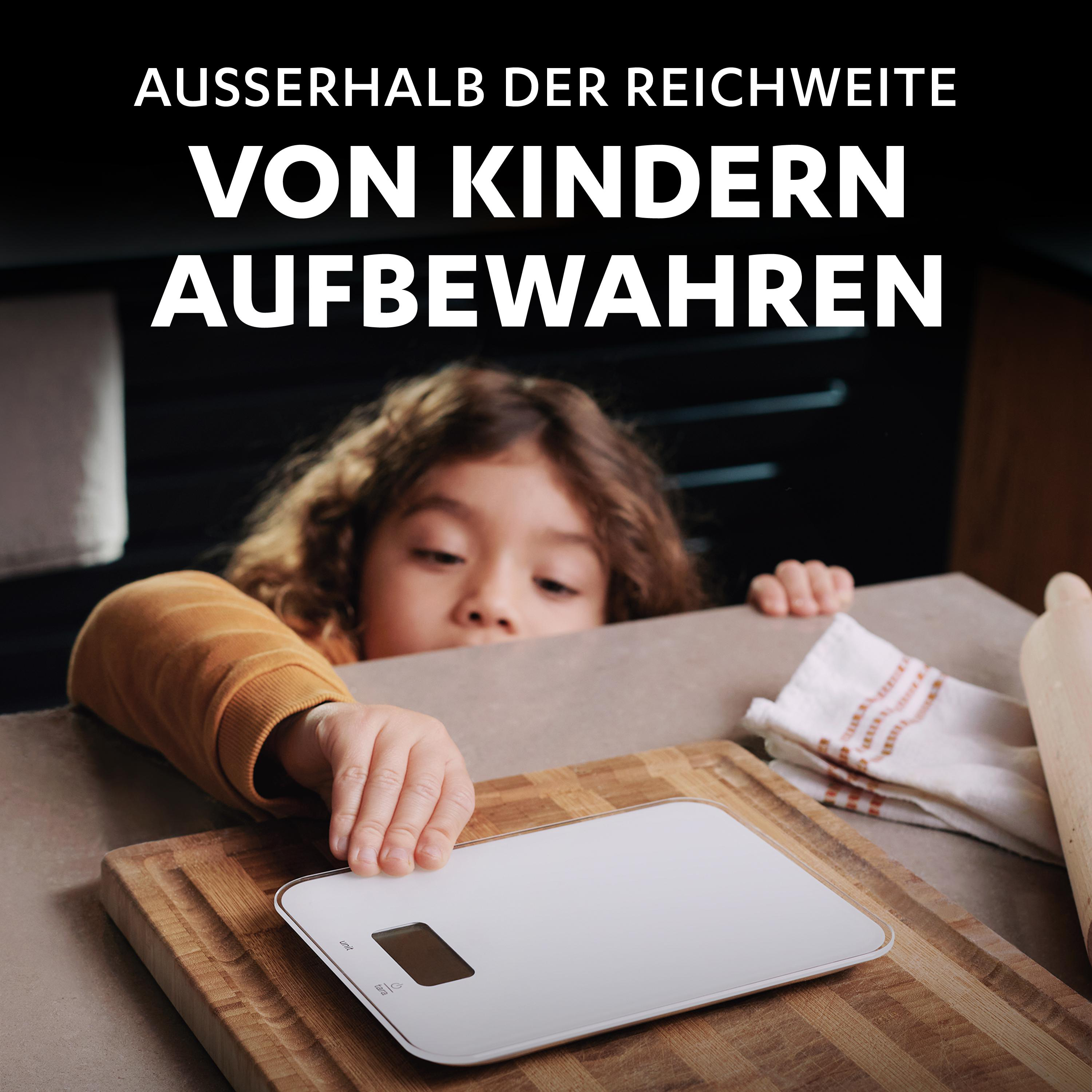 Ein Kind greift nach einer weißen Waage auf einem Holzbrett, Sicherheitswarnung oben.
