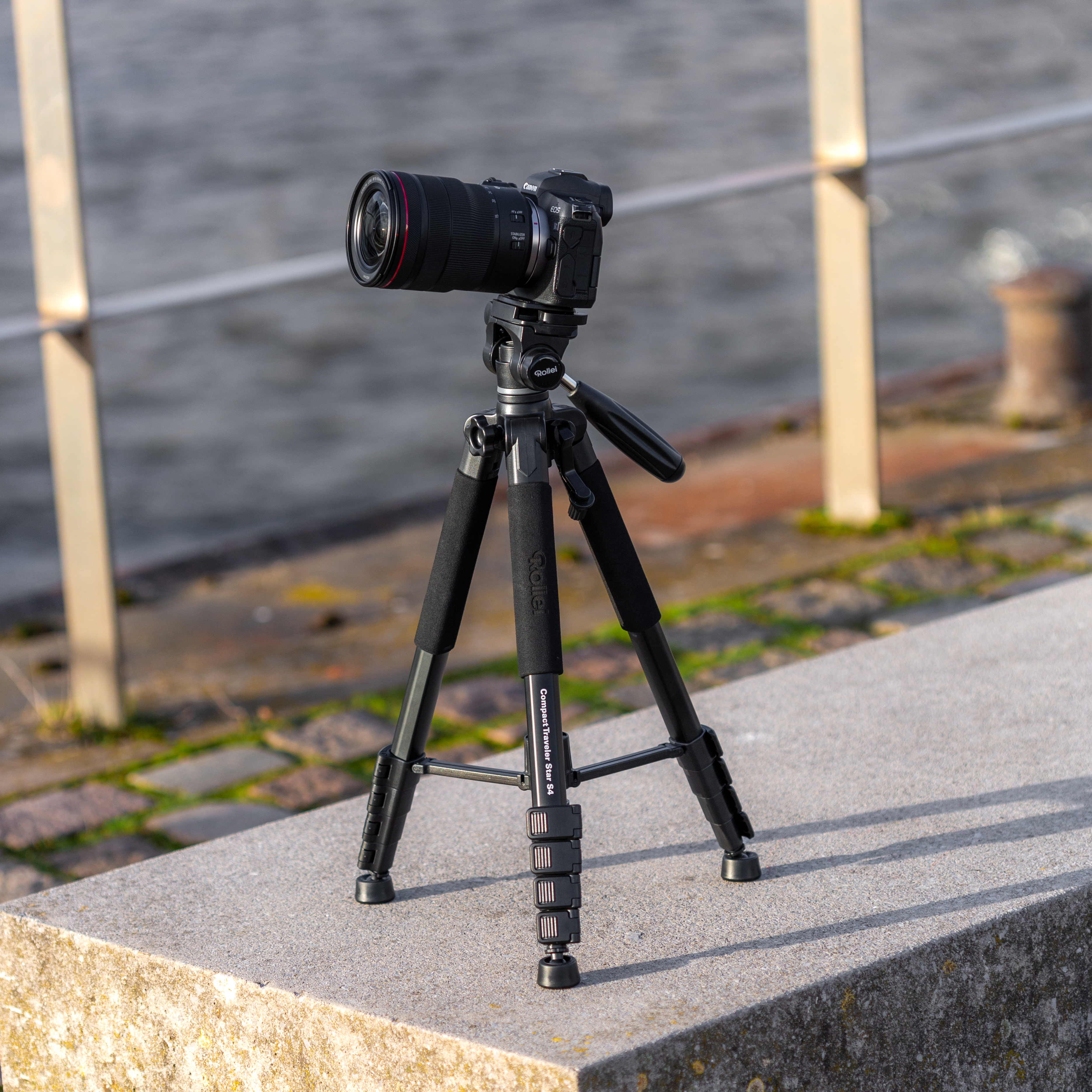 Eine schwarze Kamera auf einem Stativ steht auf Beton, im Hintergrund Wasser.