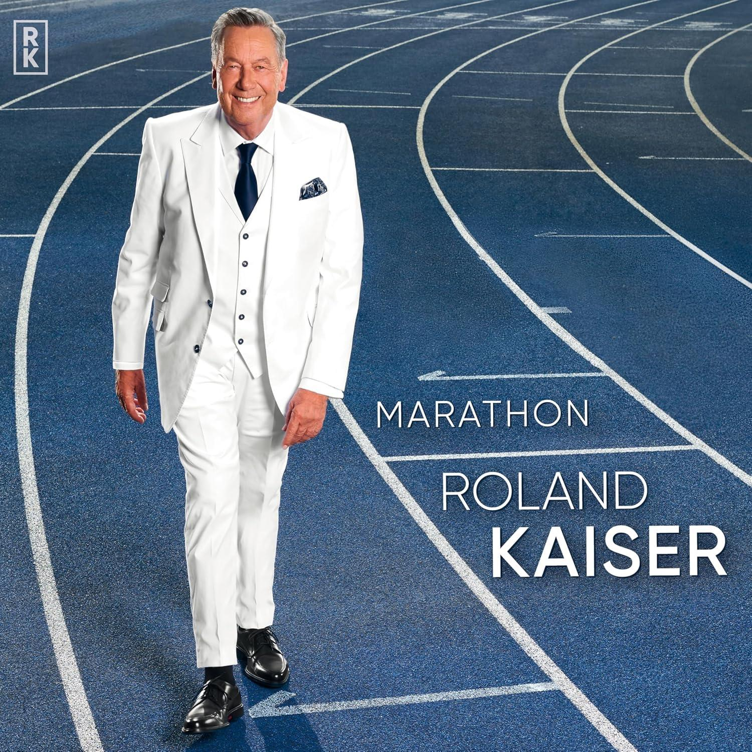 Roland Kaiser in weißem Anzug geht auf einer blauen Laufbahn. Der Text 'Roland Kaiser' ist geschrieben.