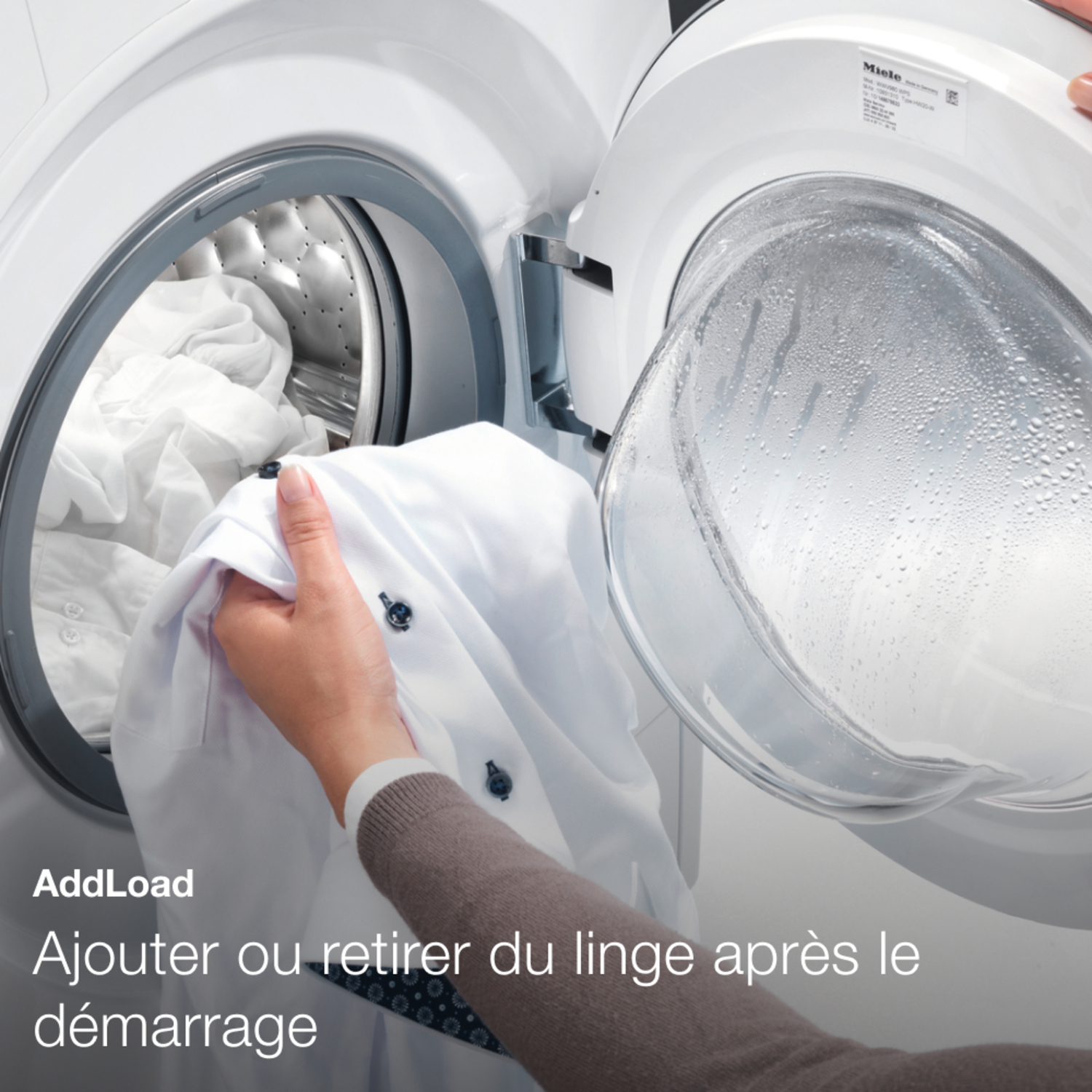 Une main met une chemise blanche dans le tambour d'une machine à laver, qui est ouvert, plein de vêtements.