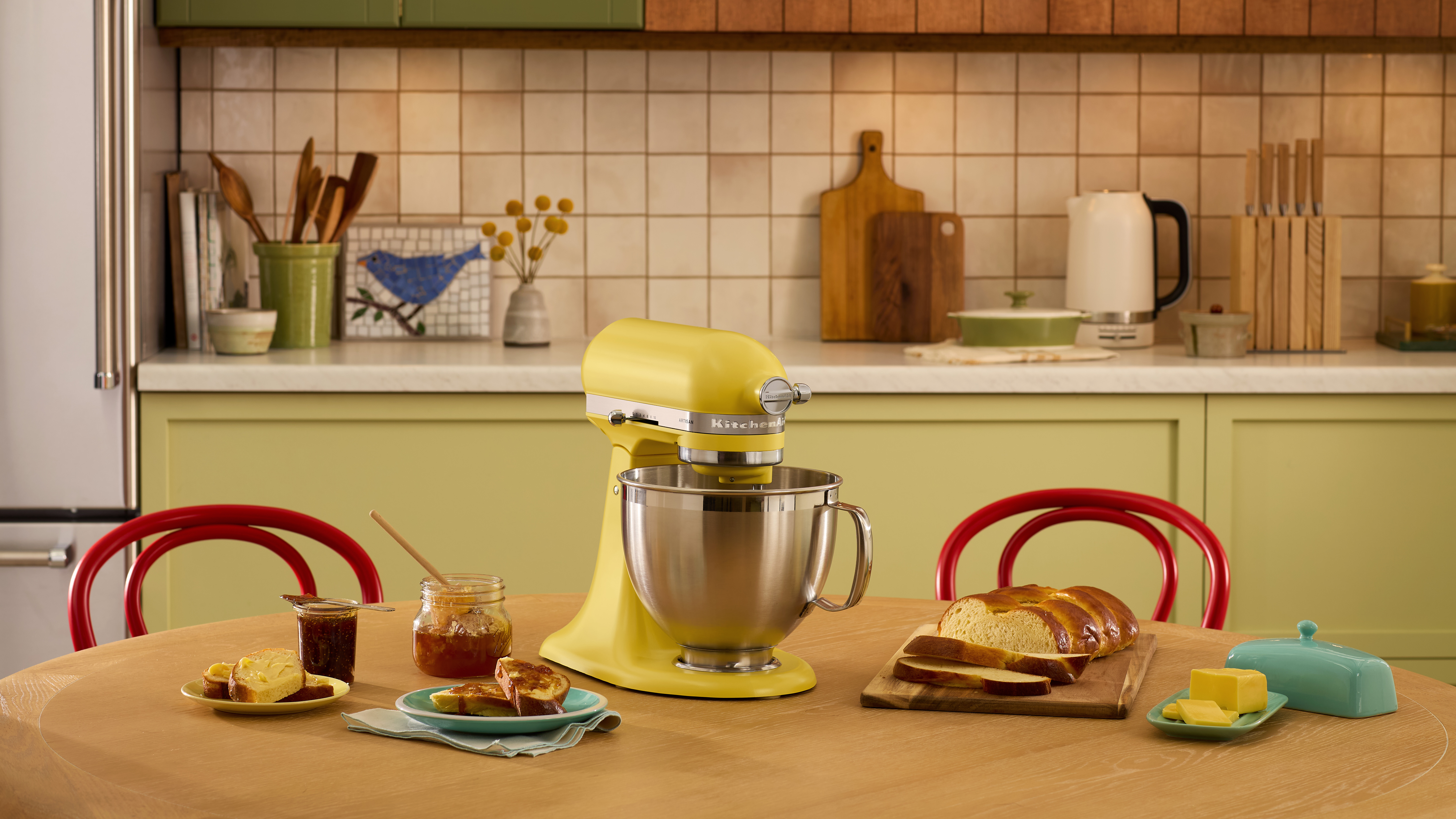 Gelber KitchenAid-Mixer auf einem Tisch mit Speisen, Schneidebrett und gemustertem Fliesenspiegel.