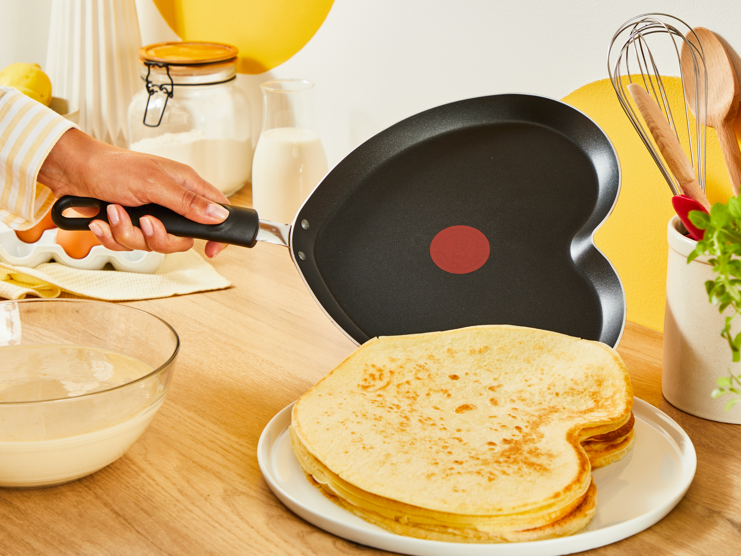Osoba trzymająca patelnię w kształcie serca z naleśnikami i składnikami na drewnianym stole.