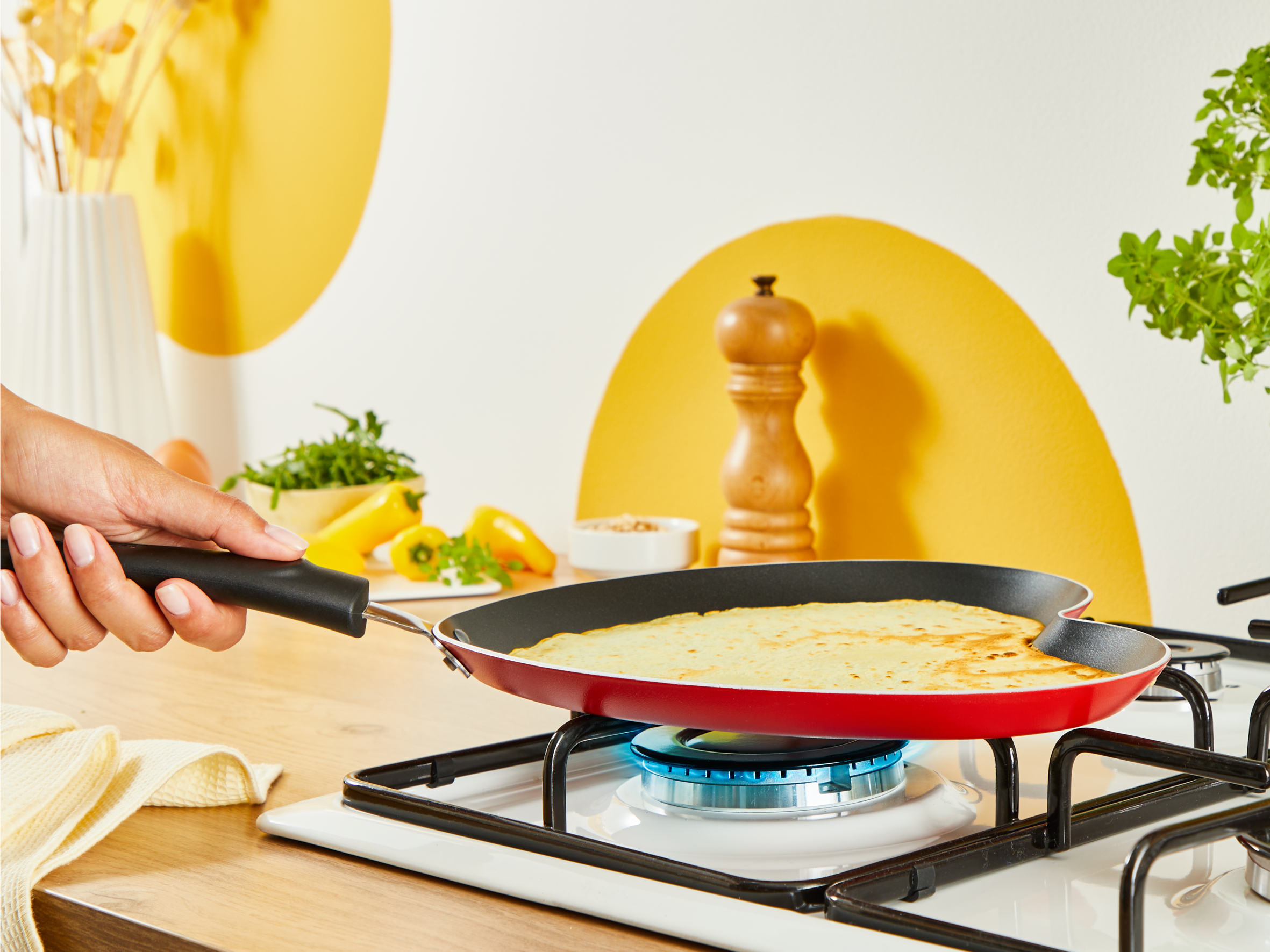 Czerwona patelnia na kuchence gazowej z naleśnikiem. Ręka trzyma uchwyt.