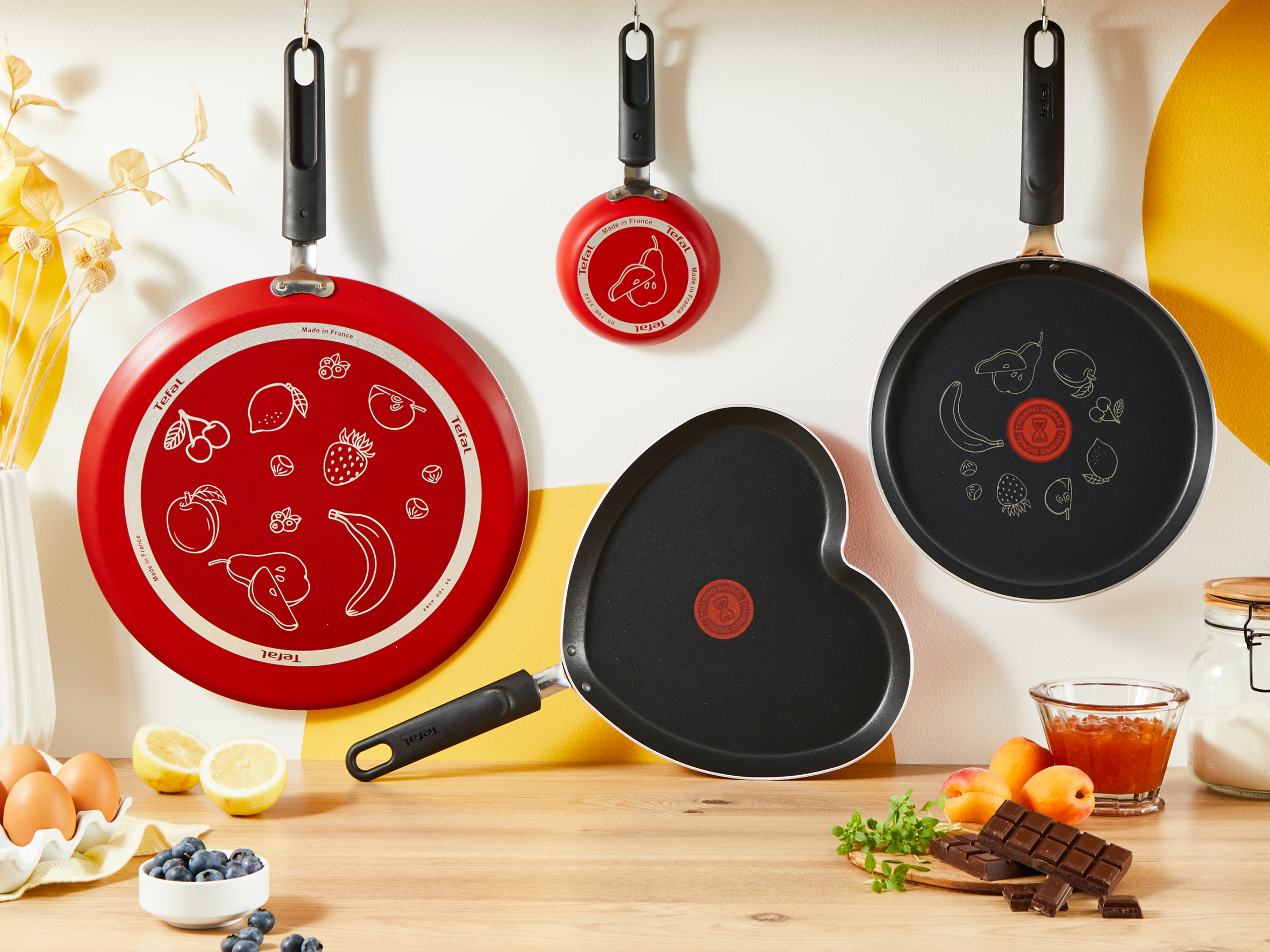 Patelnie wiszące na ścianie, w tym czerwona patelnia z ilustracją owoców.