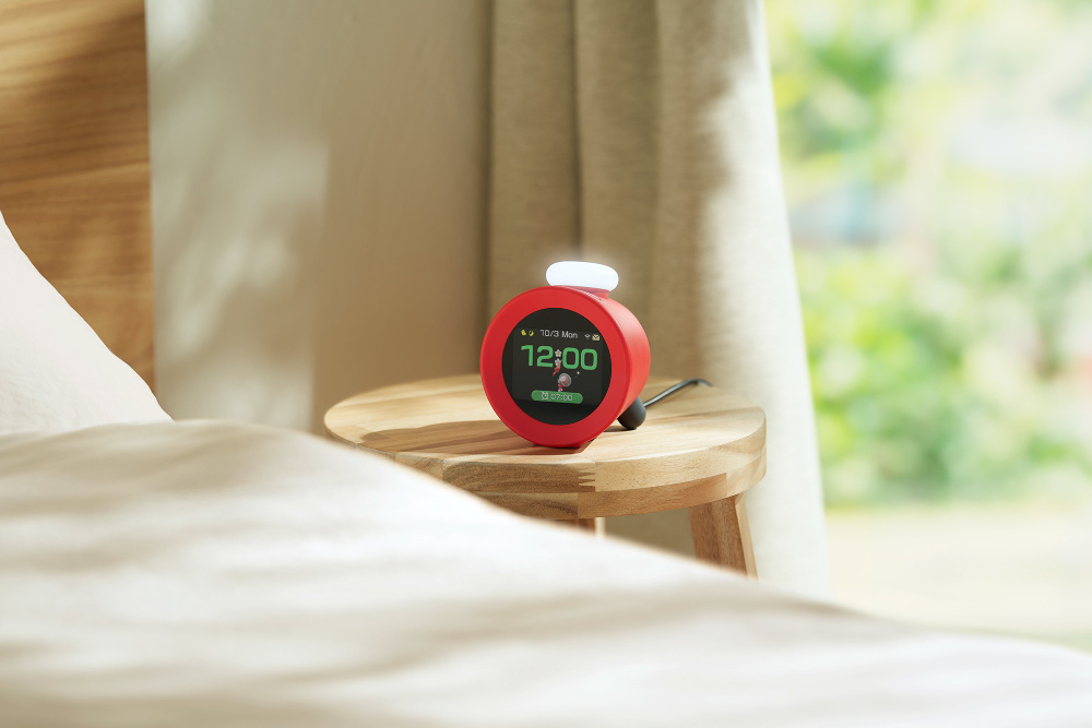 Rode wekker op een nachtkastje. Toont de tijd, 12:00. Bed en raam op de achtergrond.