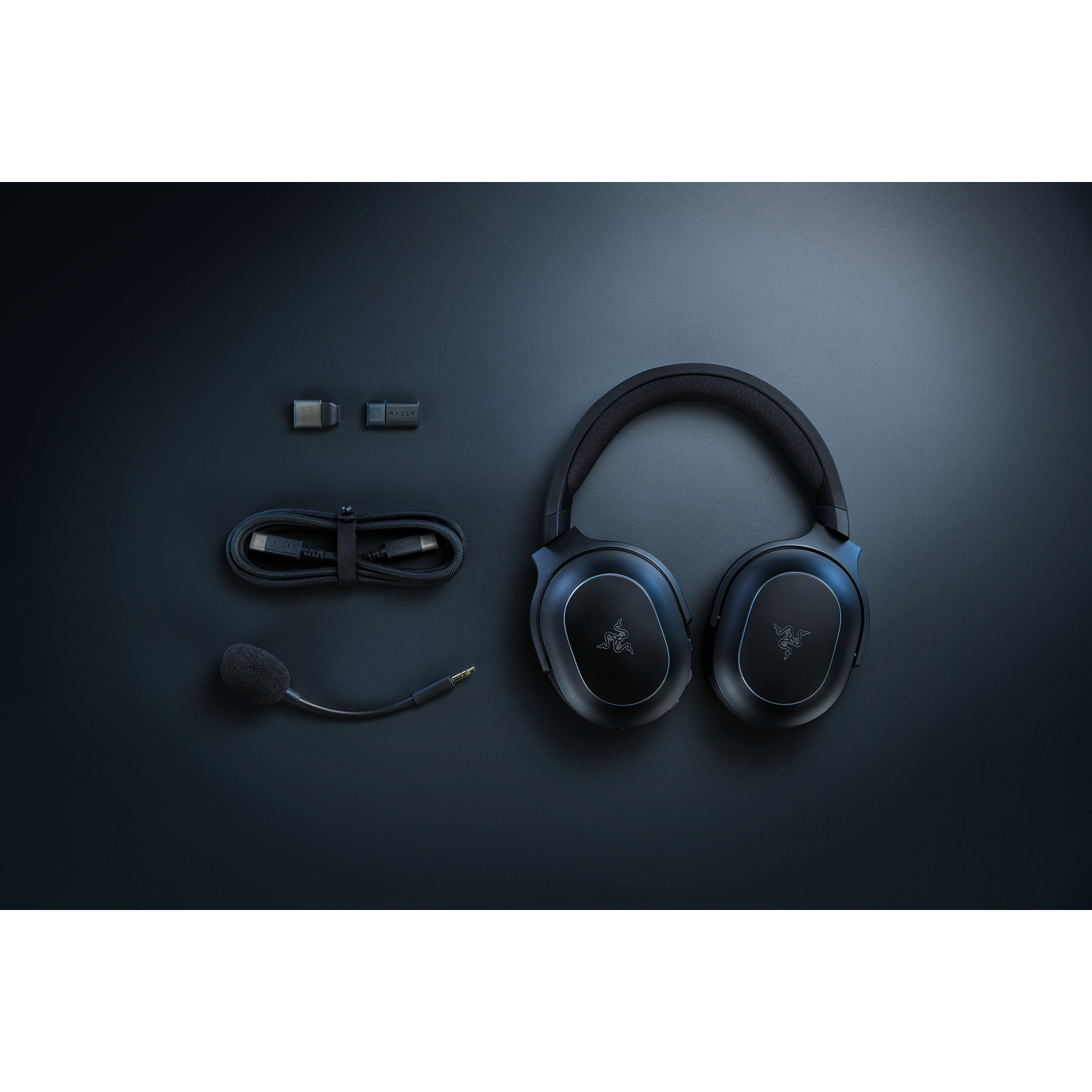 Black gaming headset and accessories on a black surface. Includes headset, mic, USB cable, and two adapters.