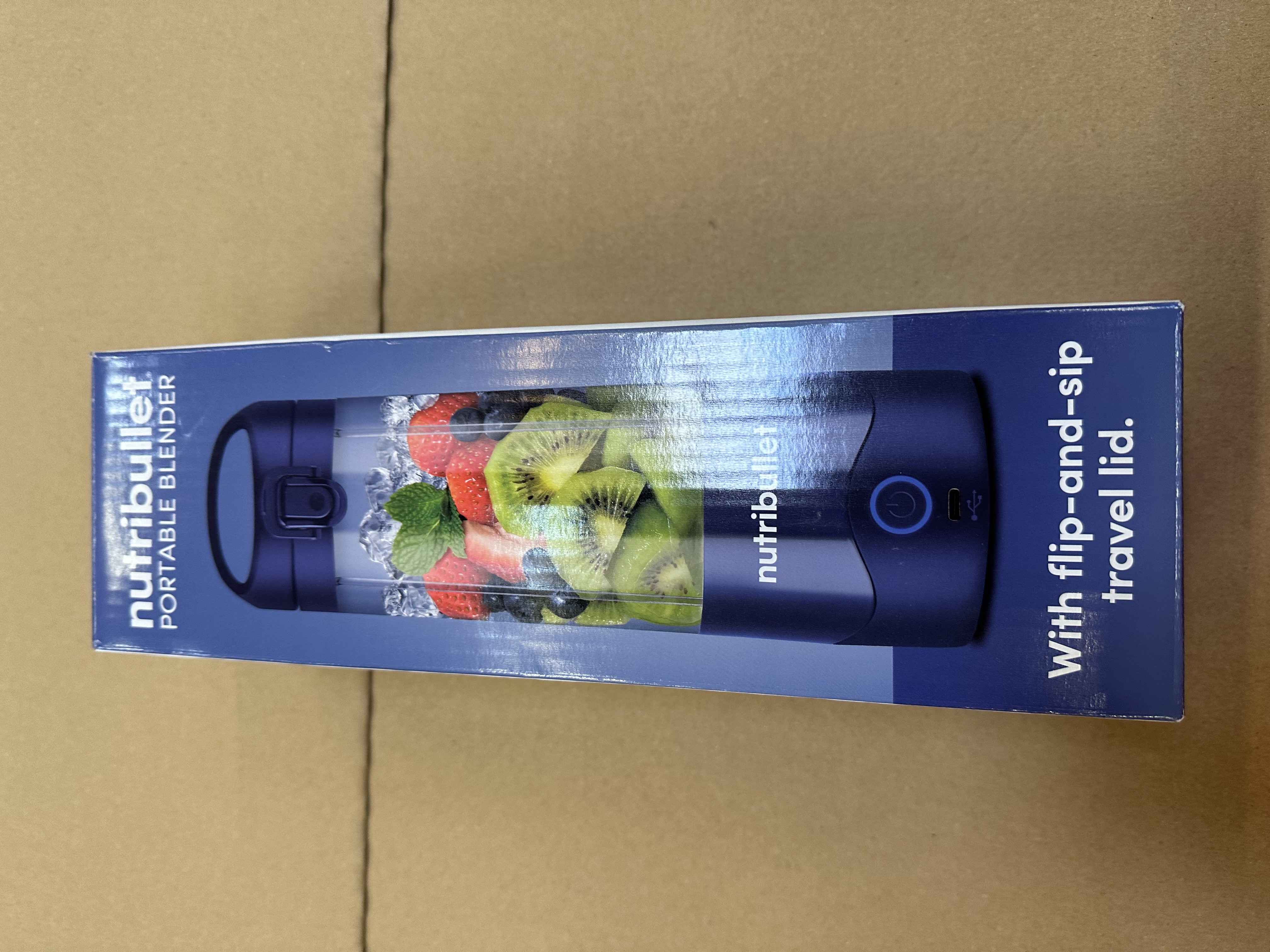 Pudełko przenośnego blendera z owocami i kostkami lodu w środku. Kolory niebieski i biały. Tekst: Nutribullet.