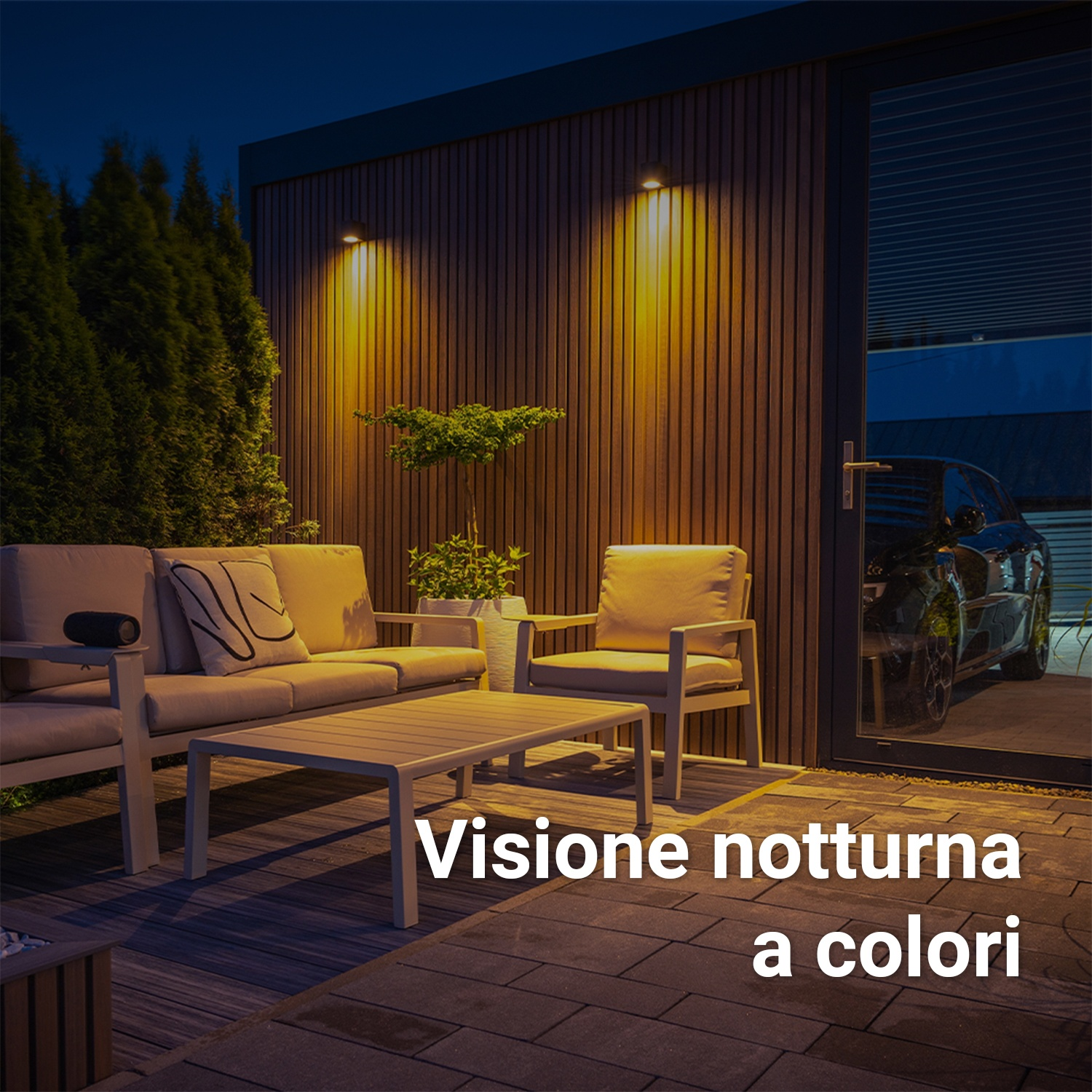 Scena all'aperto di notte con parete in legno, luci e zona salotto. Un'auto nera è parcheggiata.