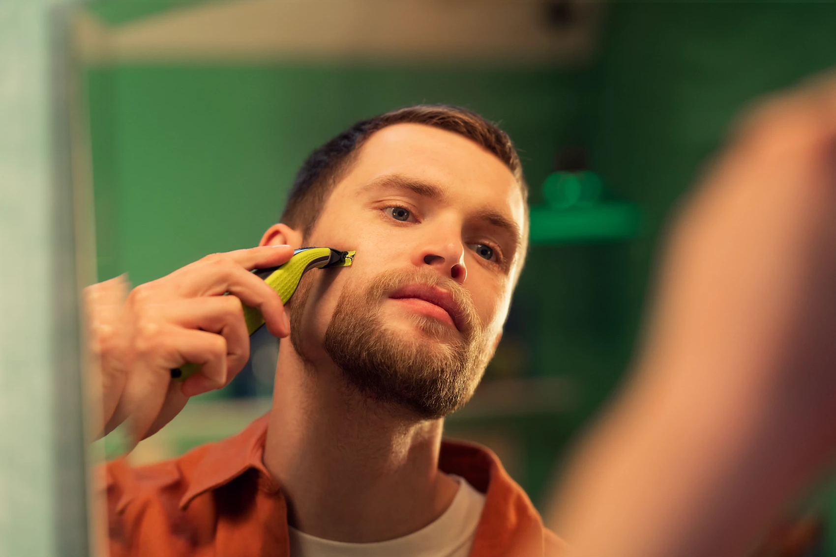 Un uomo si rade la barba con un rasoio elettrico verde e giallo in uno specchio del bagno.