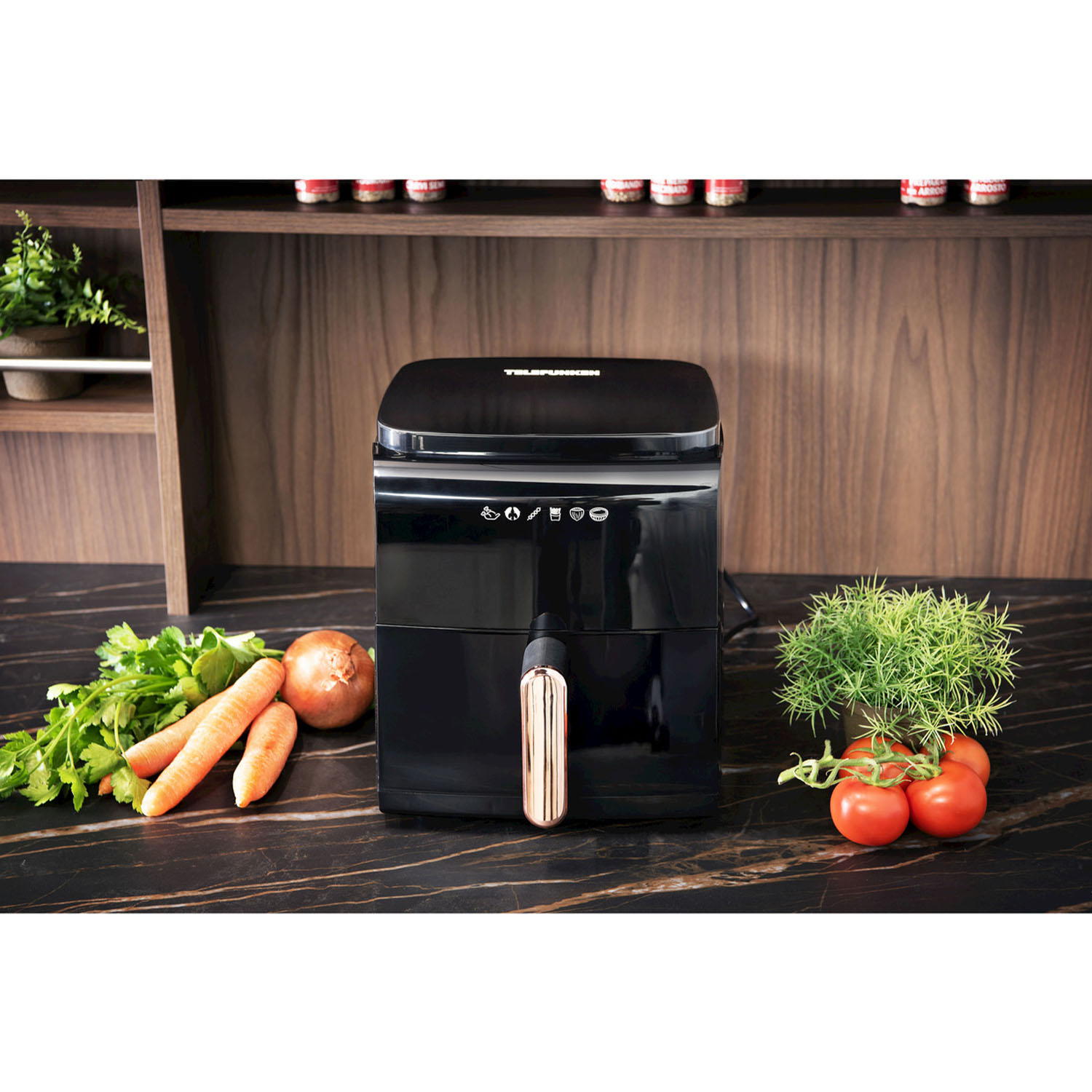 Una friggitrice ad aria nera su un piano di lavoro con verdure e pomodori. Dietro, un mobile di legno.
