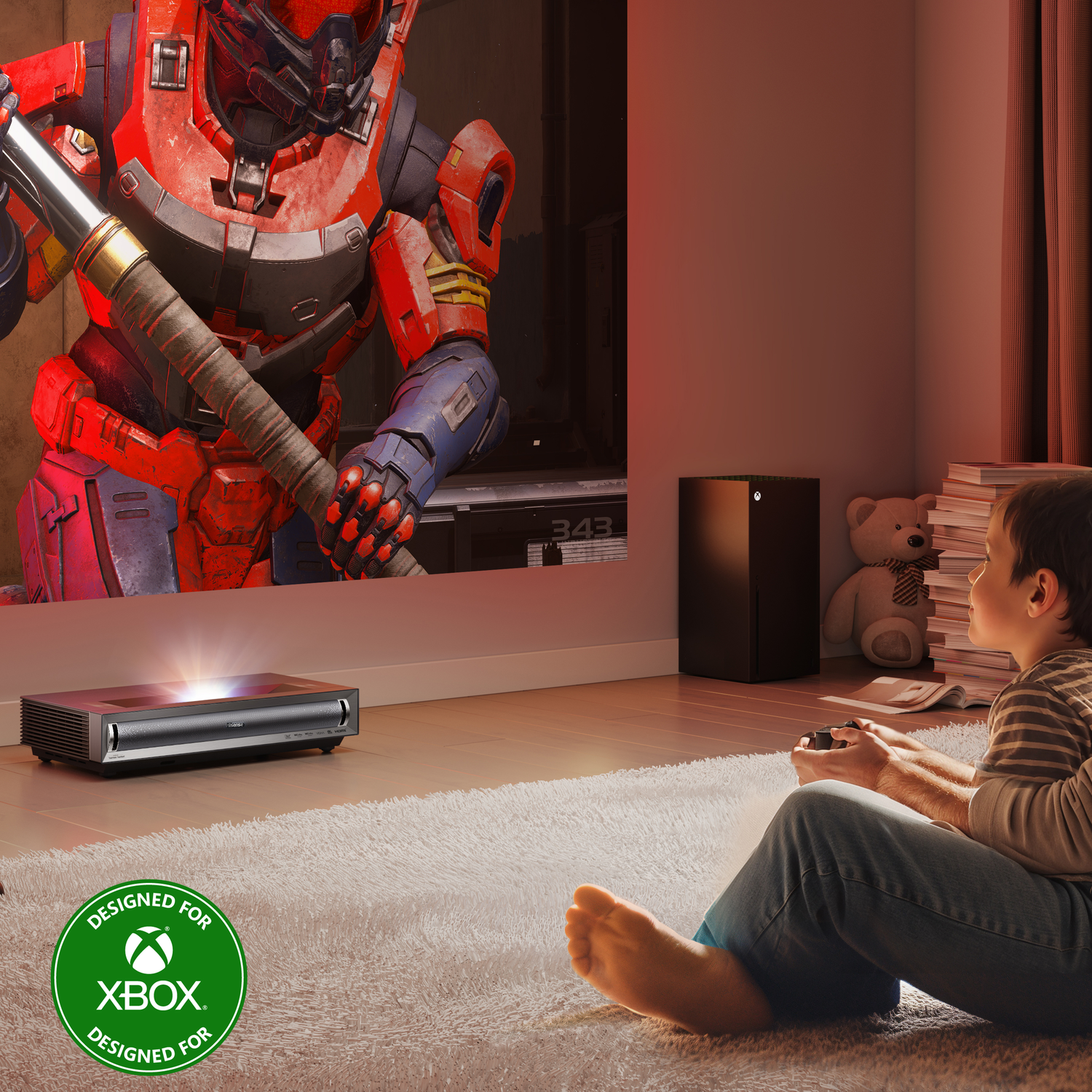 A boy plays a game projected on a wall. Projector and Xbox console are visible. Carpet and teddy bear.