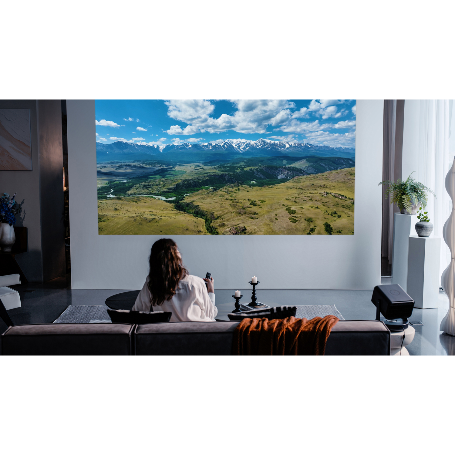 Woman watching a projected image on a wall. Snow-covered mountains and green valley are visible. Sofa.