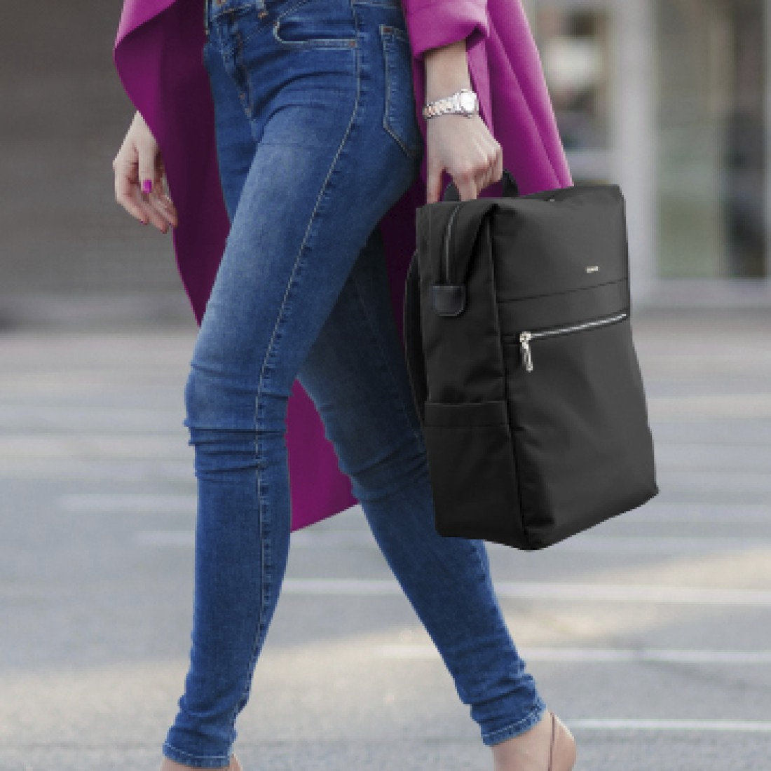 Eine Frau in einem rosa Mantel und blauen Jeans geht auf einer gepflasterten Straße und trägt einen schwarzen Rucksack.