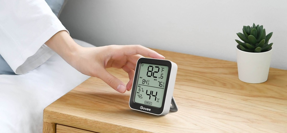Ręka osoby sięga w stronę termometru cyfrowego na drewnianym stole.