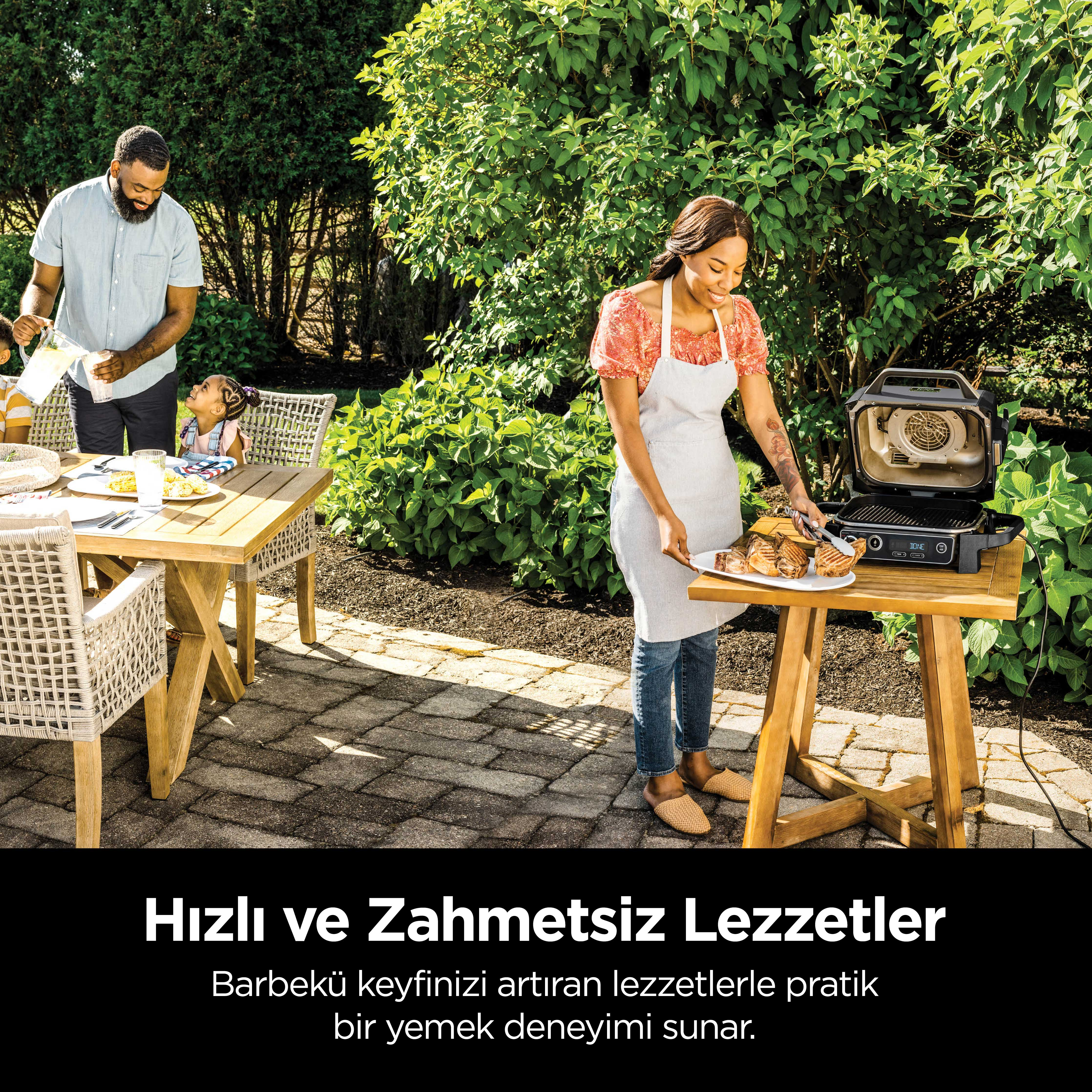 Dışarıda barbekü yapan aile. Bir kadın yemek pişiriyor, diğerleri yakındaki bir masada.