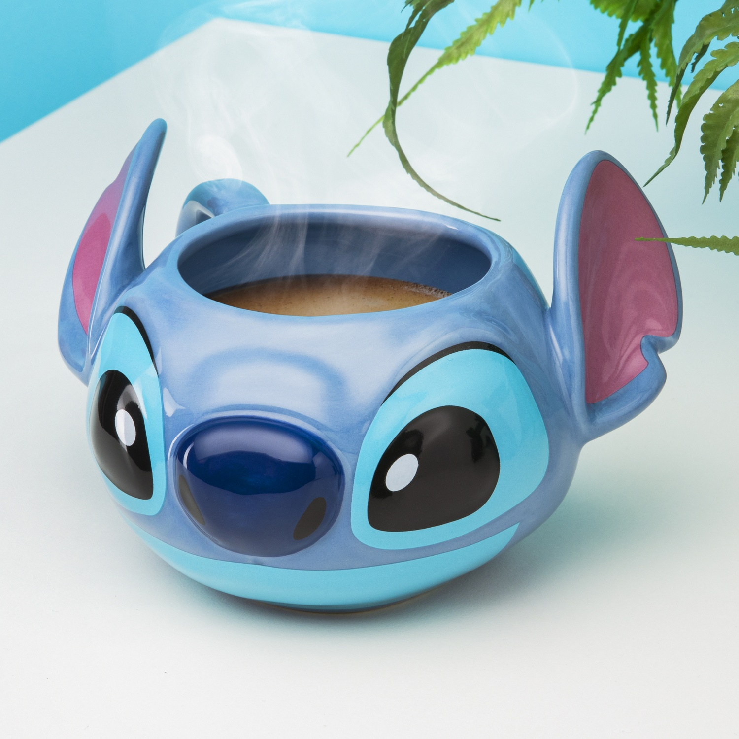 A blue Stitch mug with steam rising from coffee, on a white surface with background foliage.