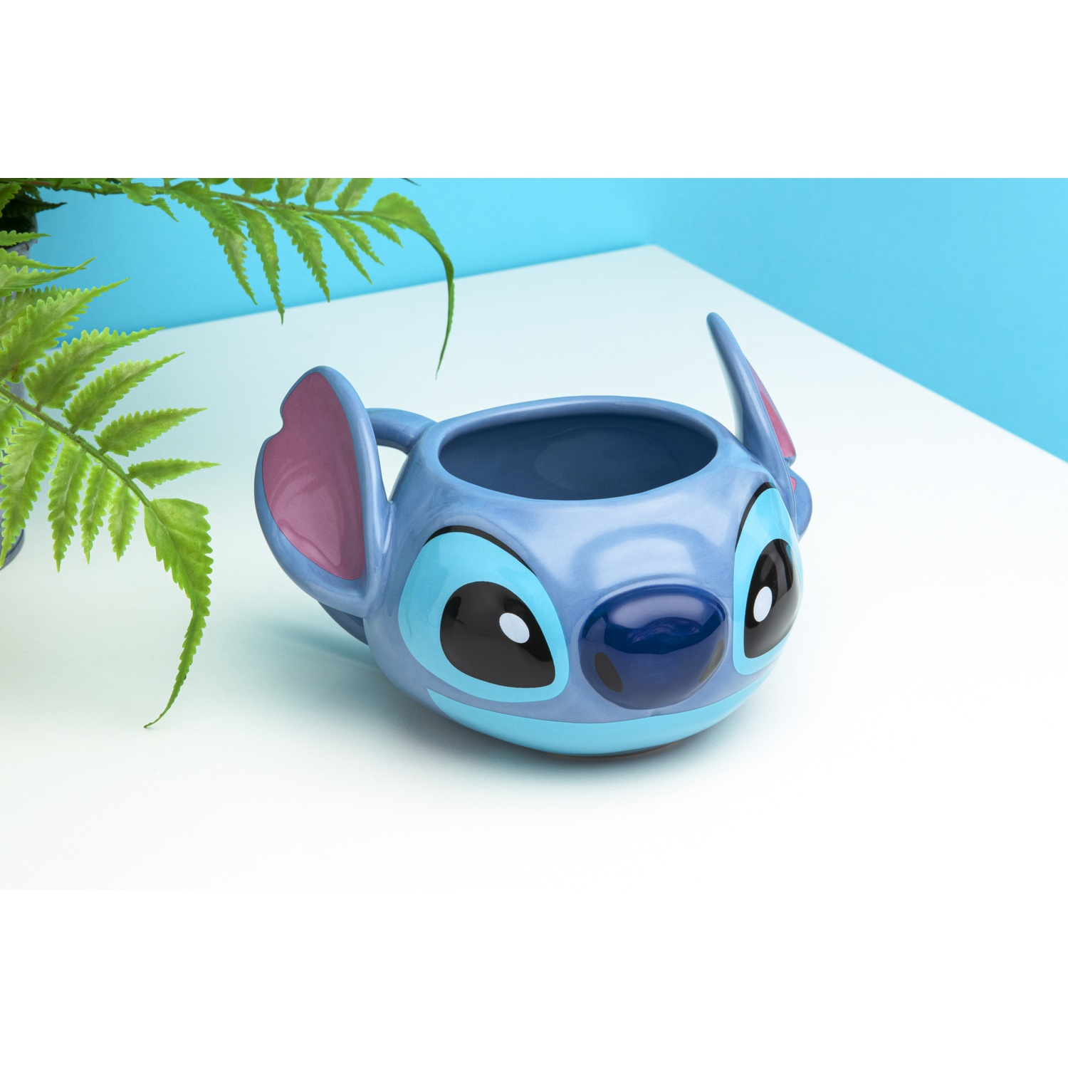 A blue Stitch mug with large ears and eyes. It is on a white surface with green plants nearby.