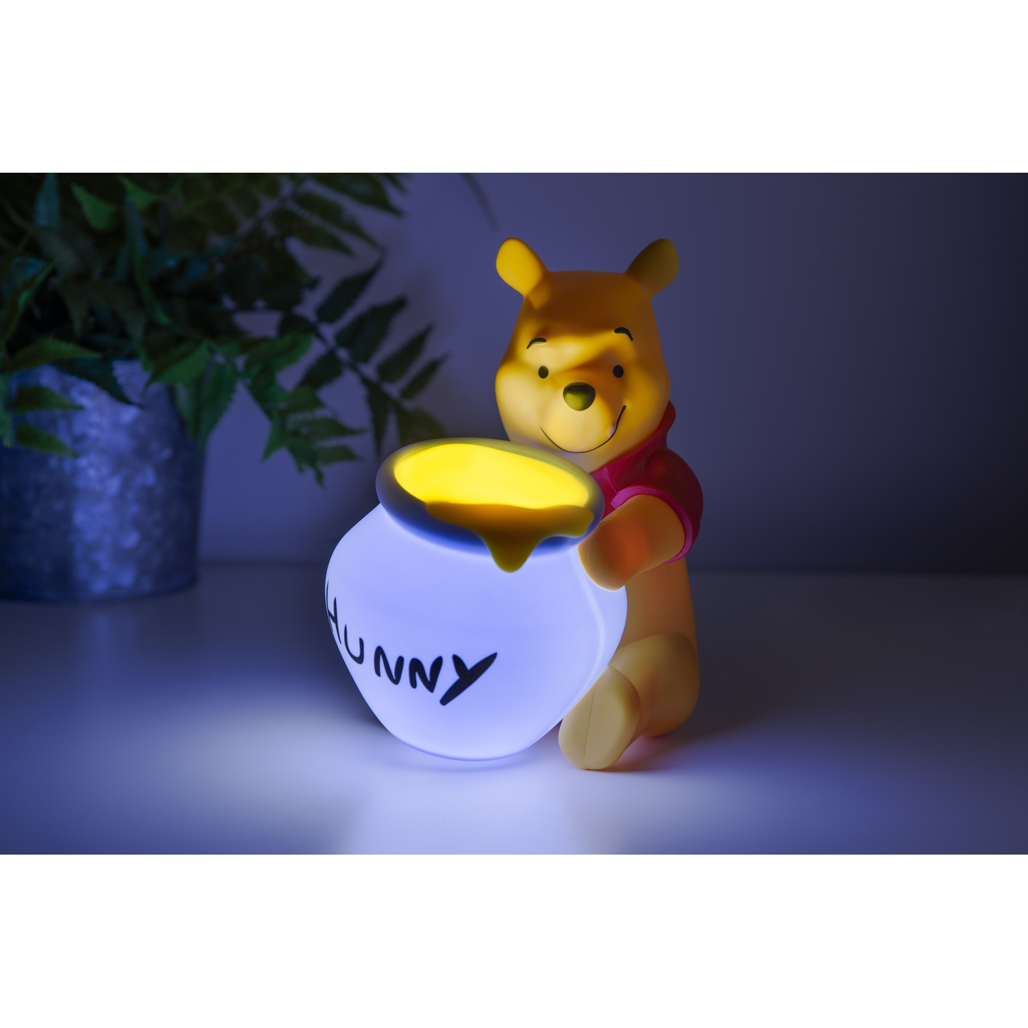 Une figurine de Winnie l'ourson avec un pot de miel brillant. Elle est éclairée dans une pièce avec une plante verte.