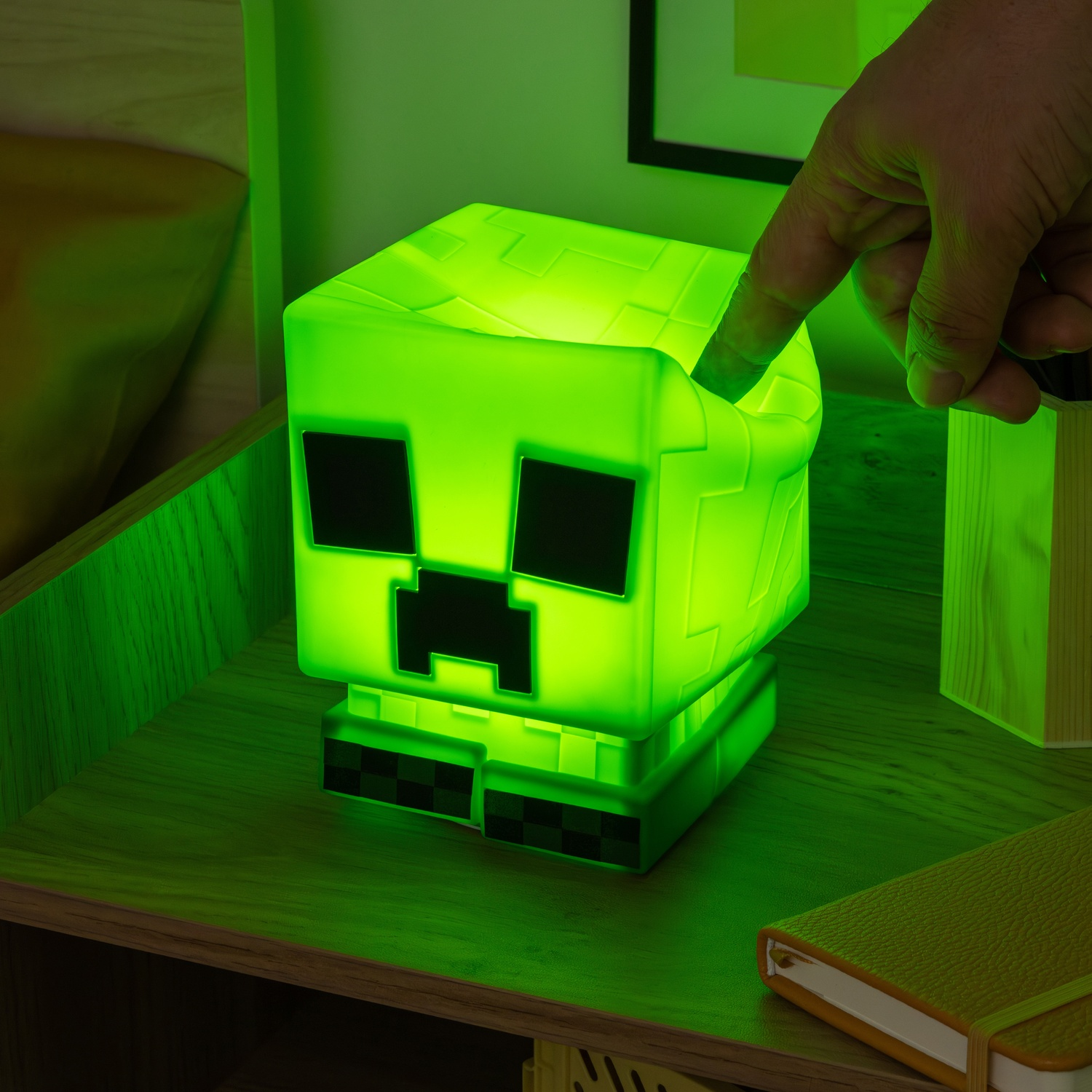Green Minecraft Creeper lamp on a nightstand. A hand is touching the lamp. There is a book nearby.