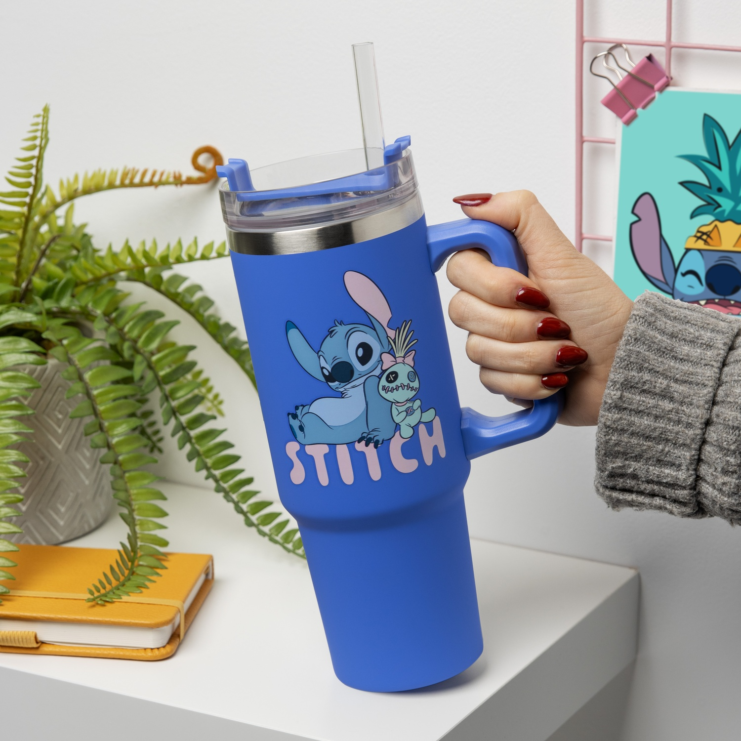 A blue Stitch tumbler is held by a hand with red nails. A Stitch drawing is in the background.