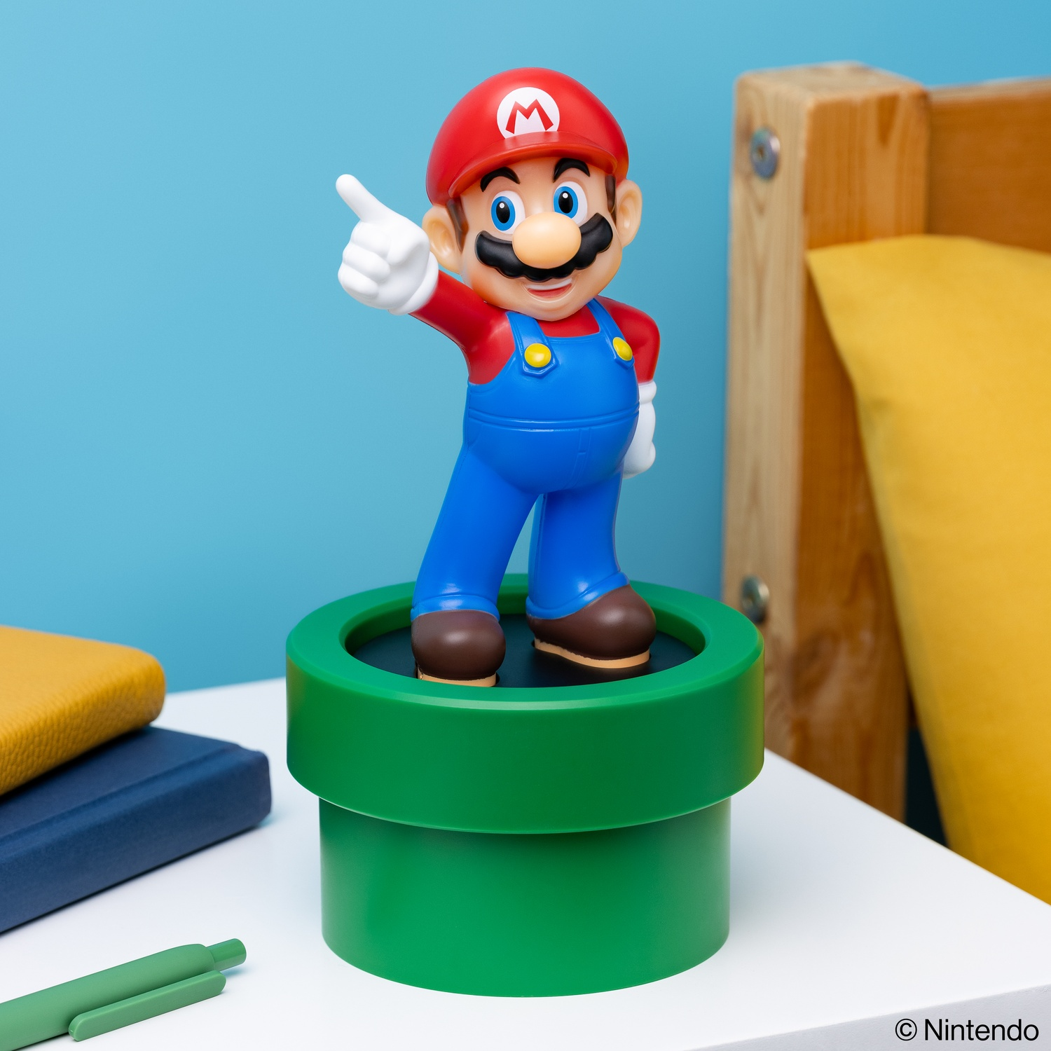 Mario figurine standing on a green pipe, pointing, on a white table, with blue and yellow background.