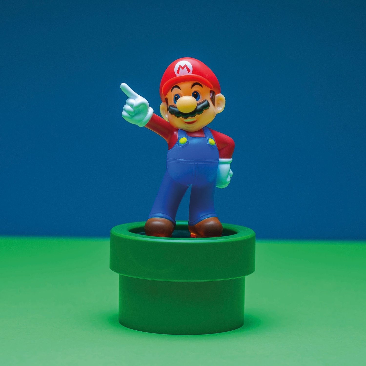 Mario figurine on a green pipe, blue and green background. Mario is pointing with his right hand.