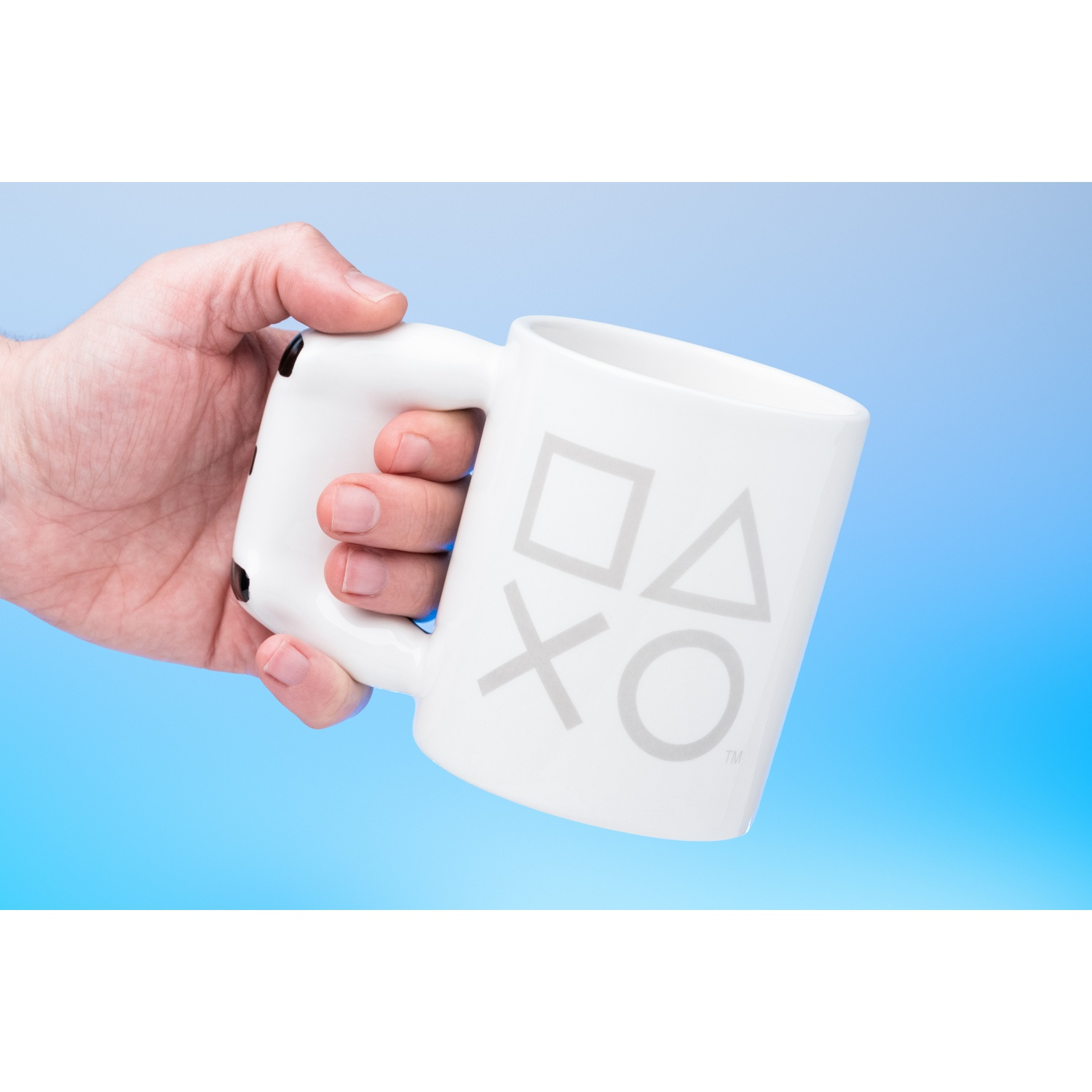 White mug with PlayStation symbols held by a hand against a blue background.