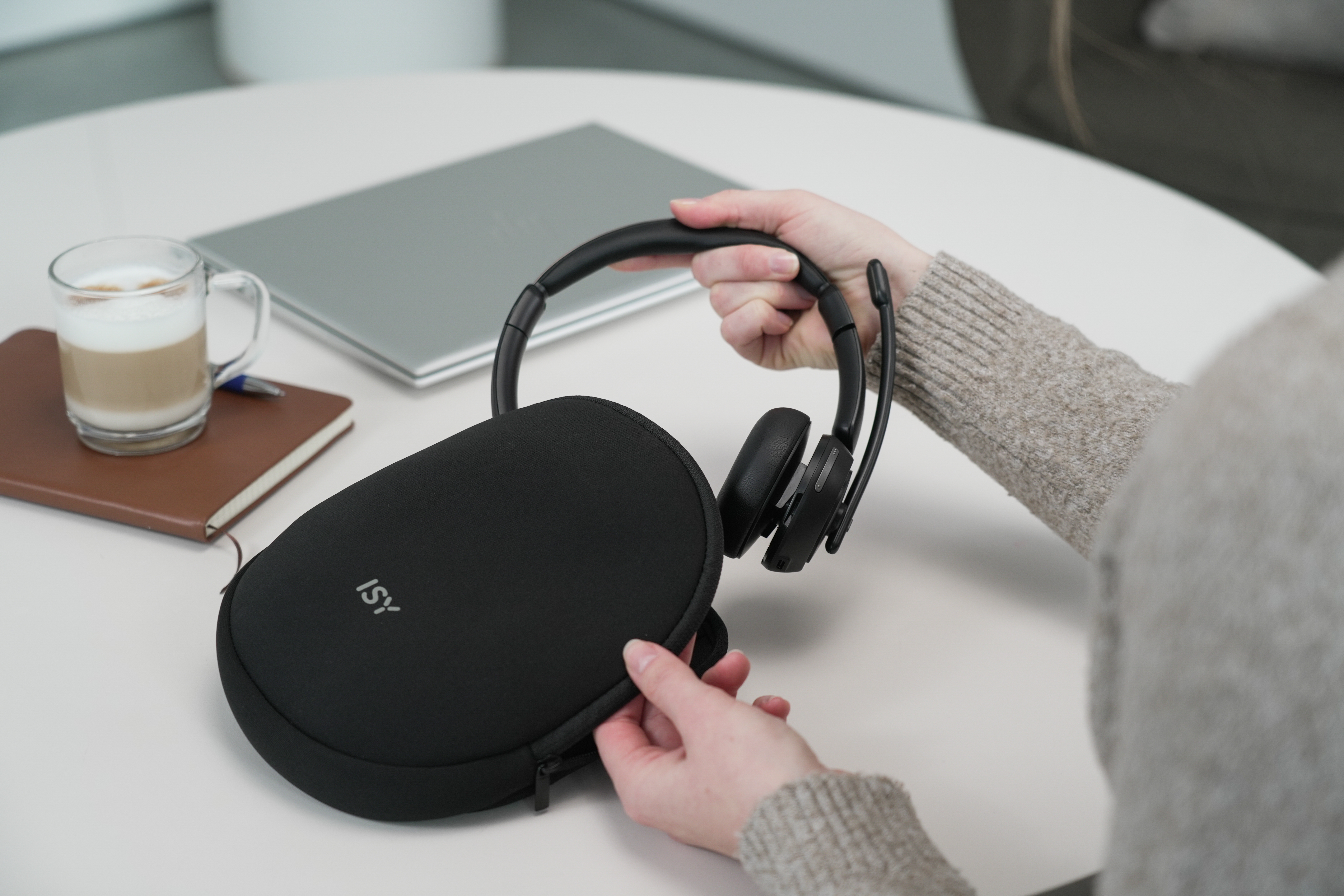 Eine Person, die ein schwarzes Headset und eine Hülle auf einem weißen Tisch mit einem Laptop und Kaffee hält.