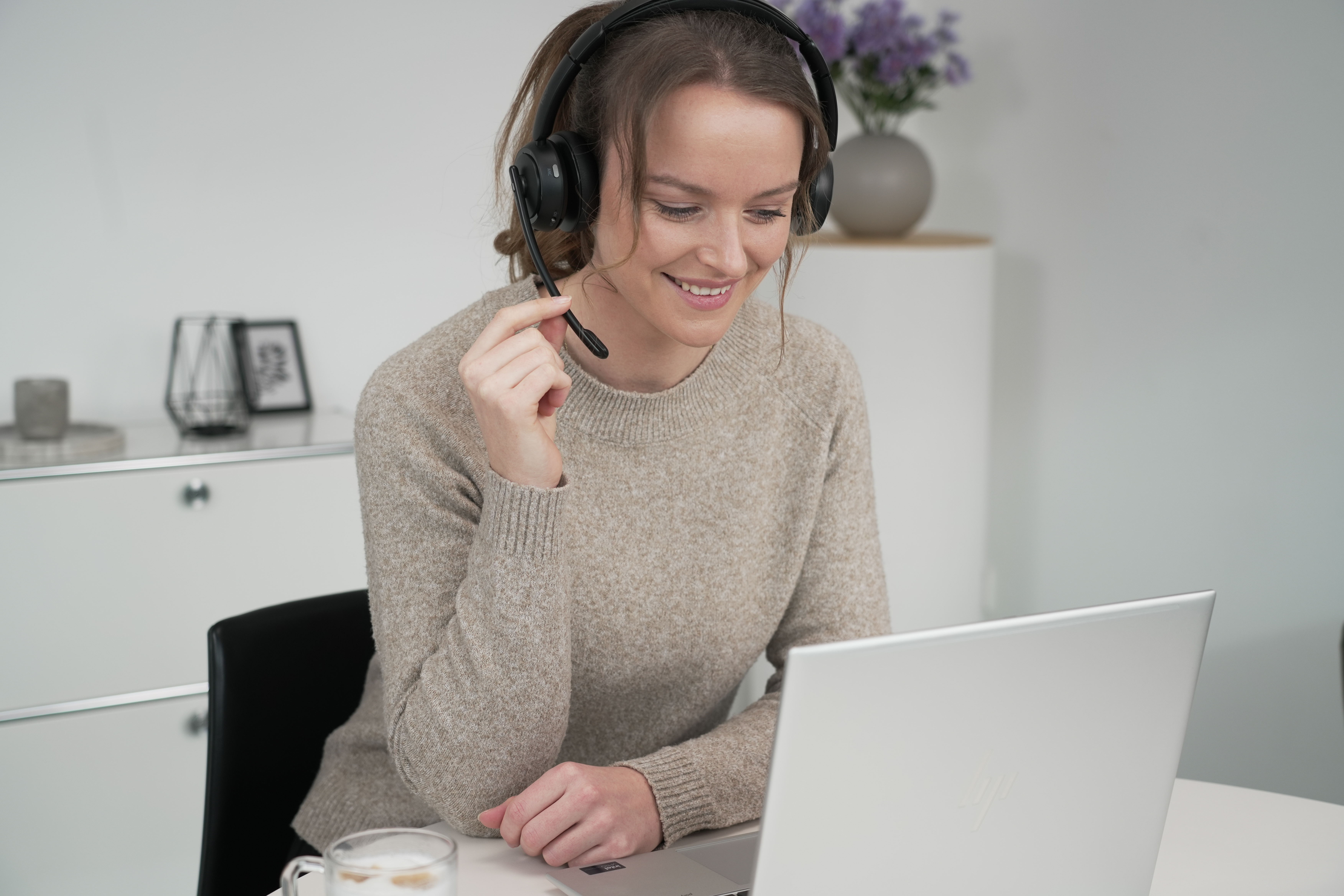 Eine lächelnde Frau mit Headset, die einen Laptop benutzt, mit einem Glas Flüssigkeit auf dem Tisch.