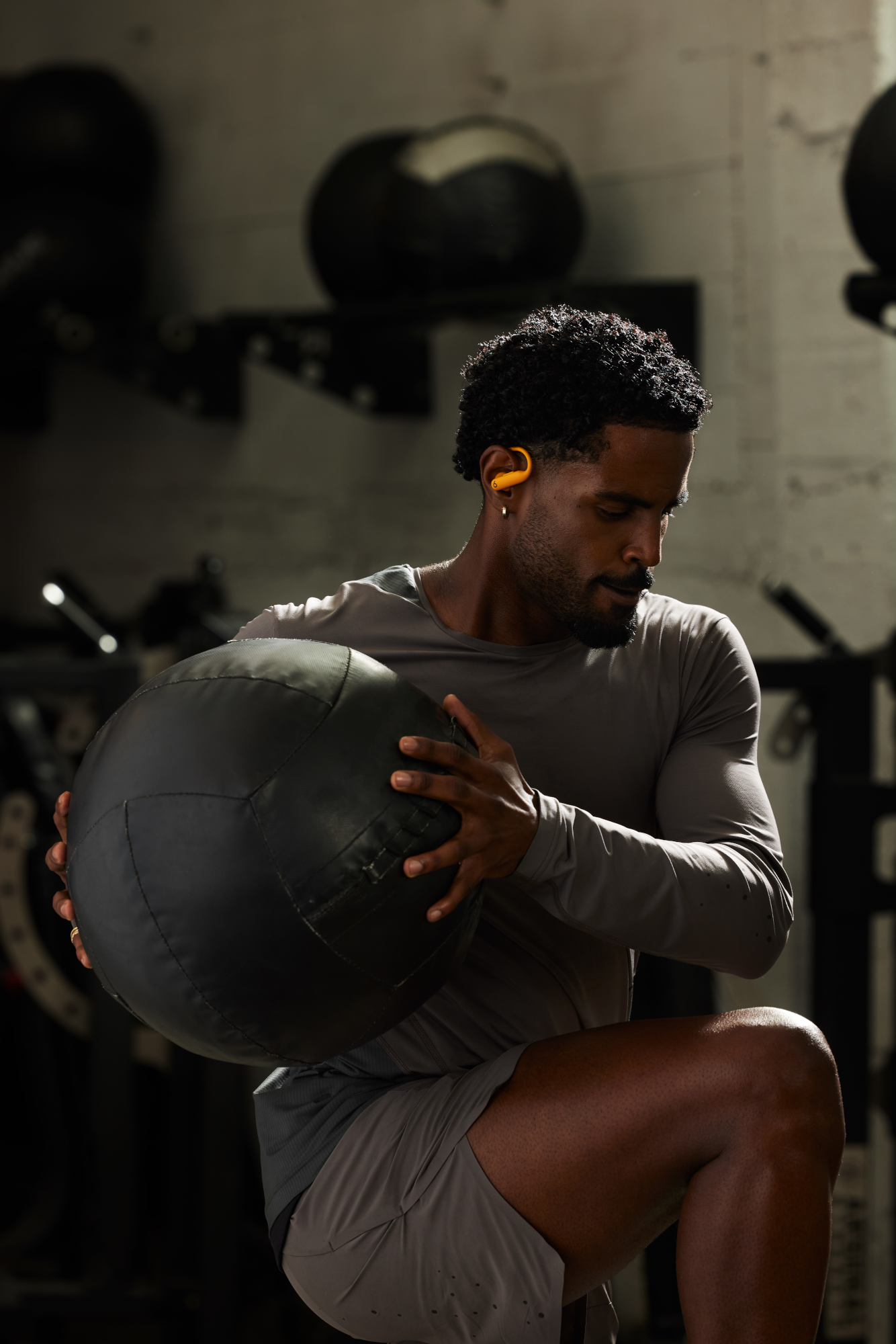 Homme dans une salle de sport tenant un ballon médicinal noir. Chemise et short gris. Écouteur jaune.