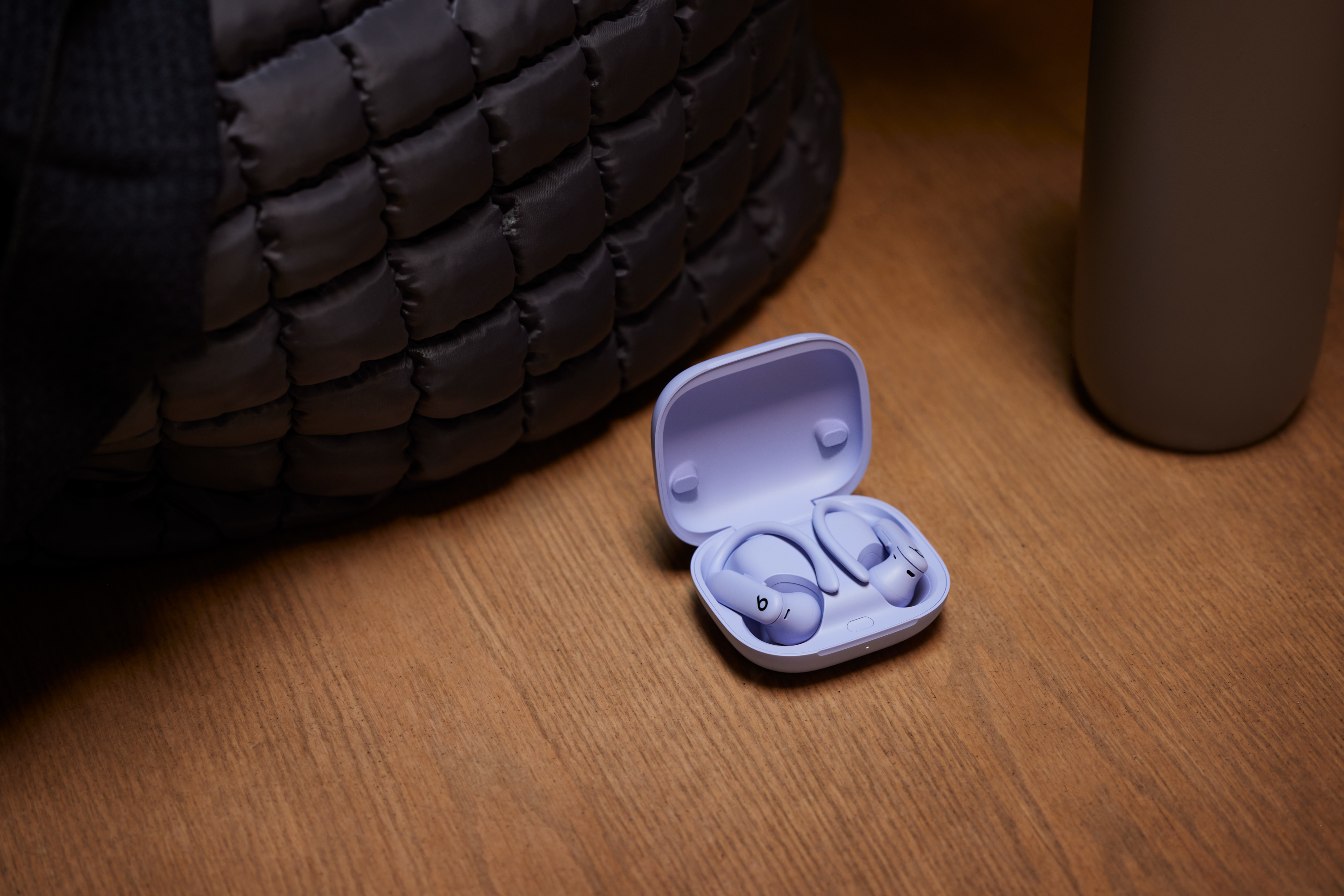Écouteurs et étui Beats violets, sur une surface en bois à côté d'un sac noir et d'un gobelet gris.