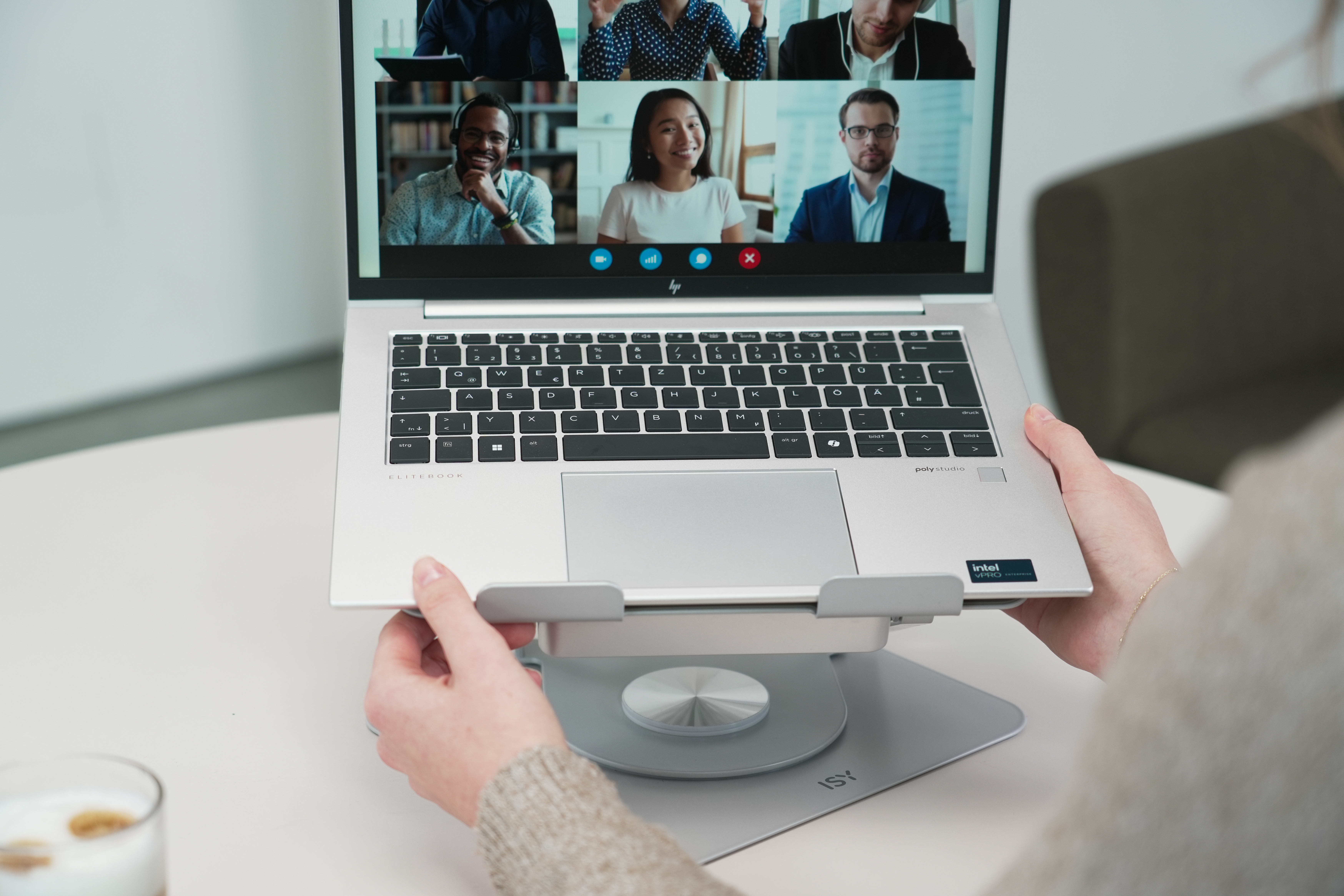 Hände halten Laptop auf einem Ständer, Videokonferenz auf dem Bildschirm. Weißer Tisch, unscharfer Hintergrund.