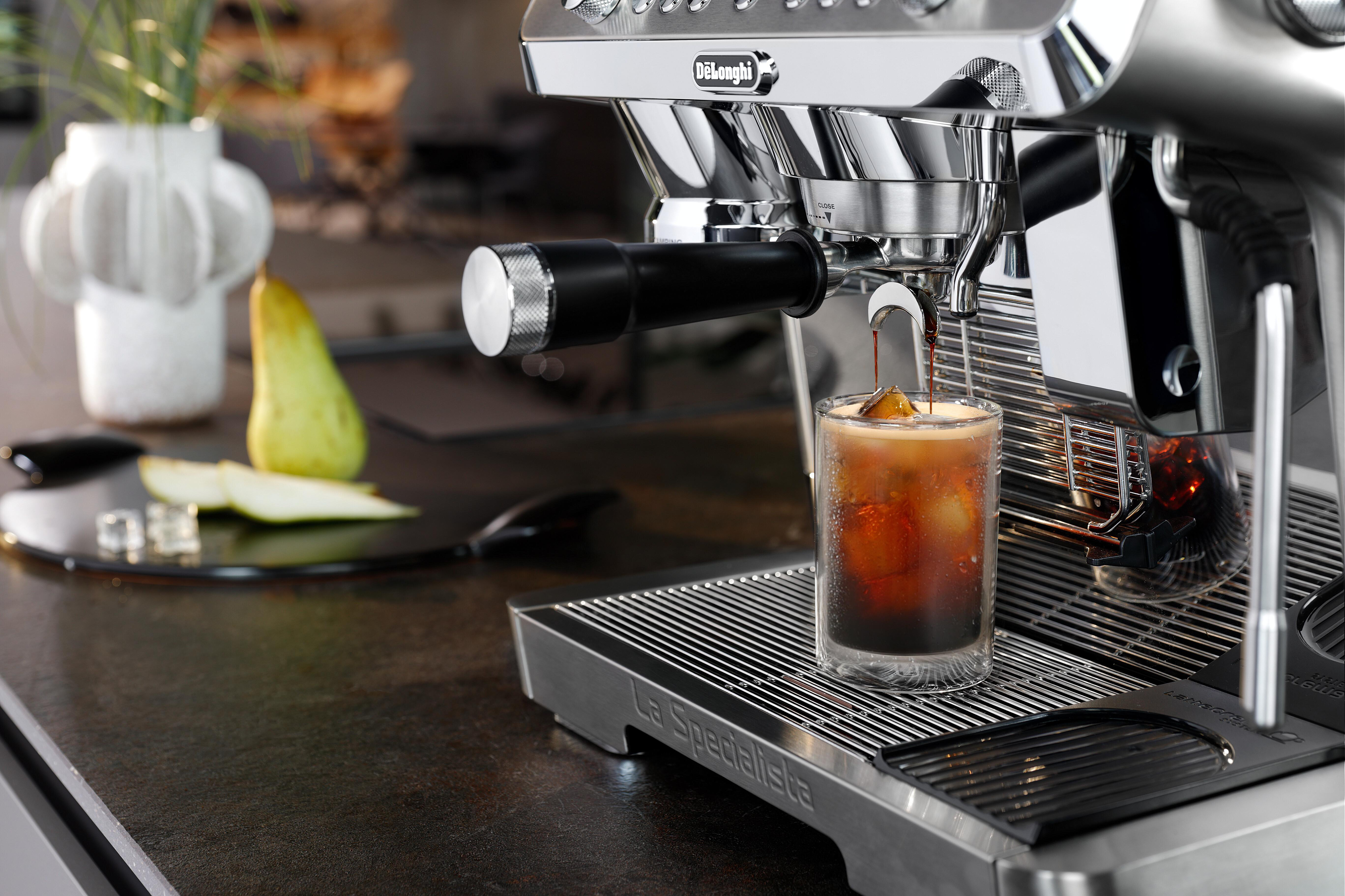 Espressomaschine gießt Kaffee in ein Glas mit Eiswürfeln. Birnenscheiben und eine Vase sind im Hintergrund.