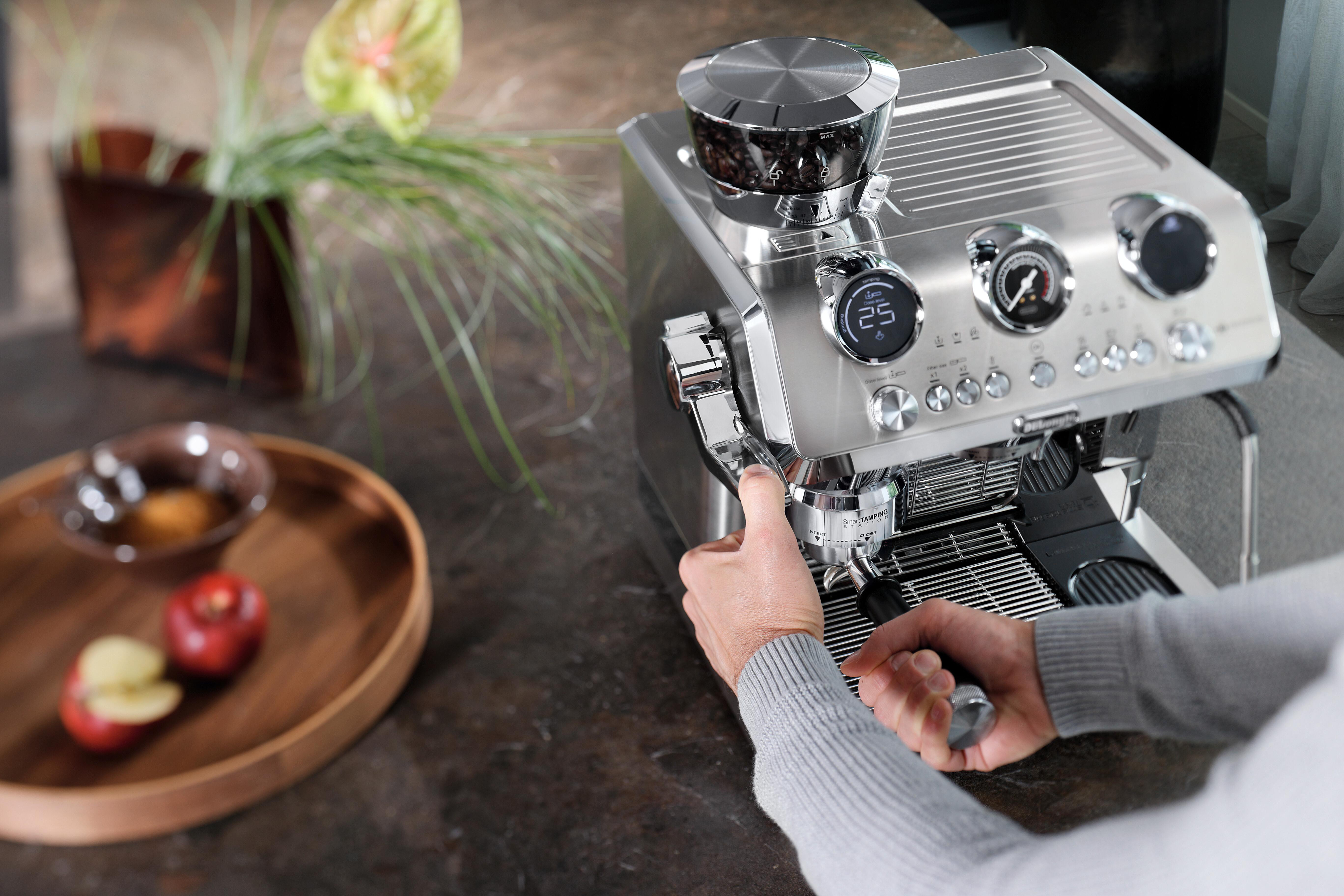 Eine Person bedient eine silberne Espressomaschine. Kaffeebohnen befinden sich in einer Mühle. Äpfel und eine Schüssel stehen daneben.