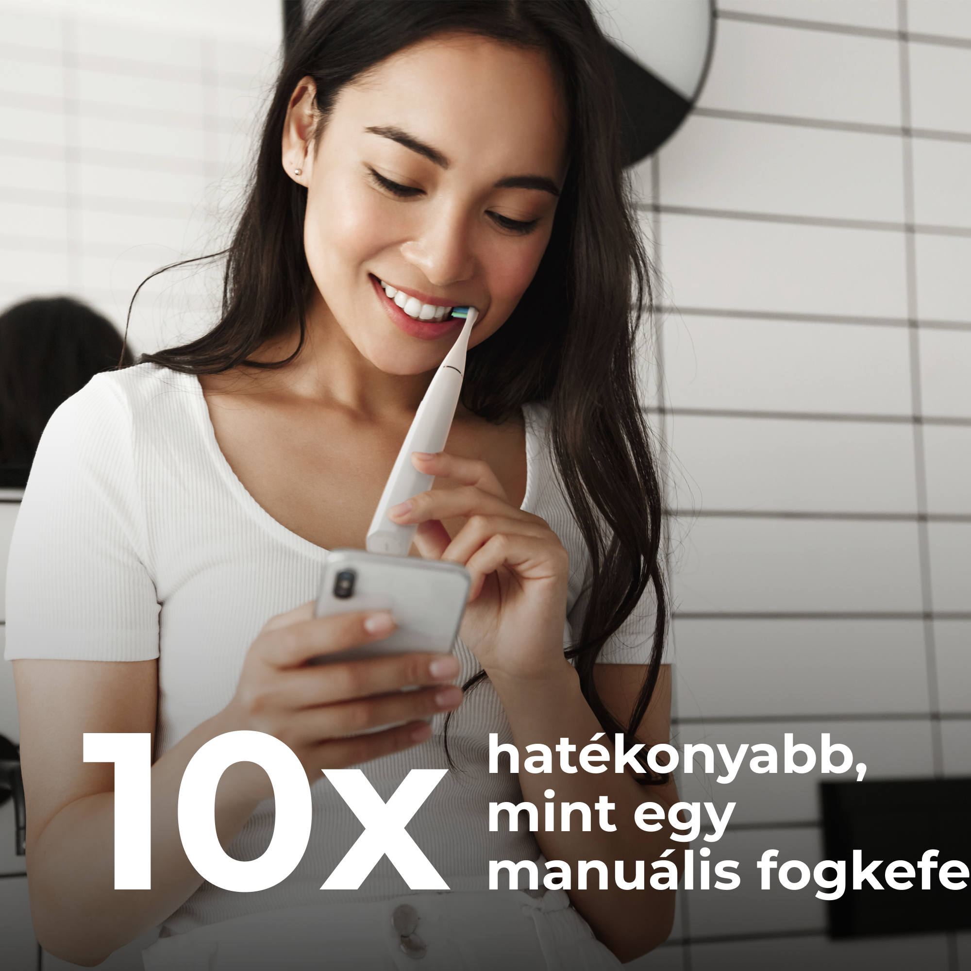 Mosolygó nő elektromos fogkefével tisztítja a fogát, miközben a telefonját nézi.