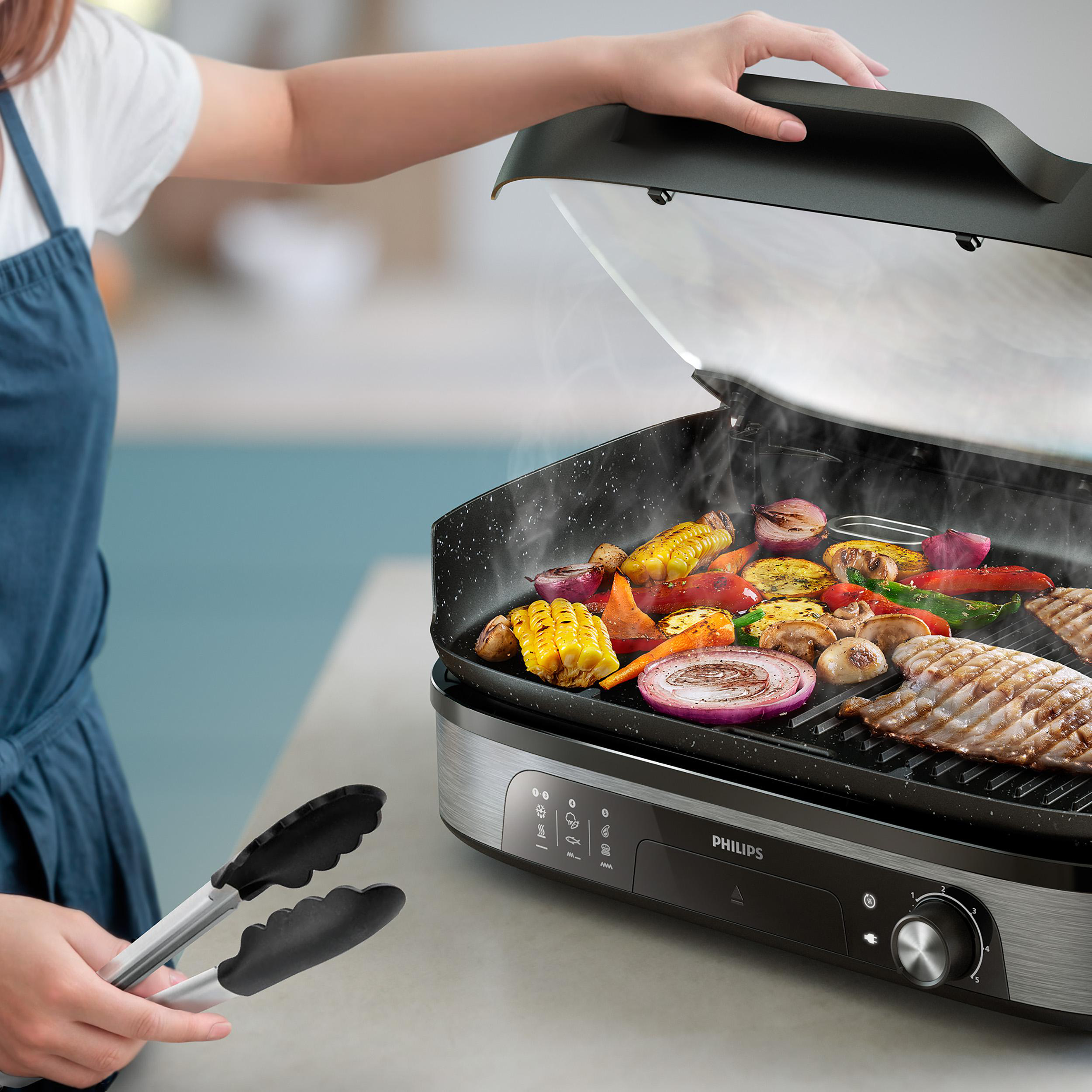 Eine Person grillt Gemüse und Steak auf einem Elektrogrill mit Zangen.