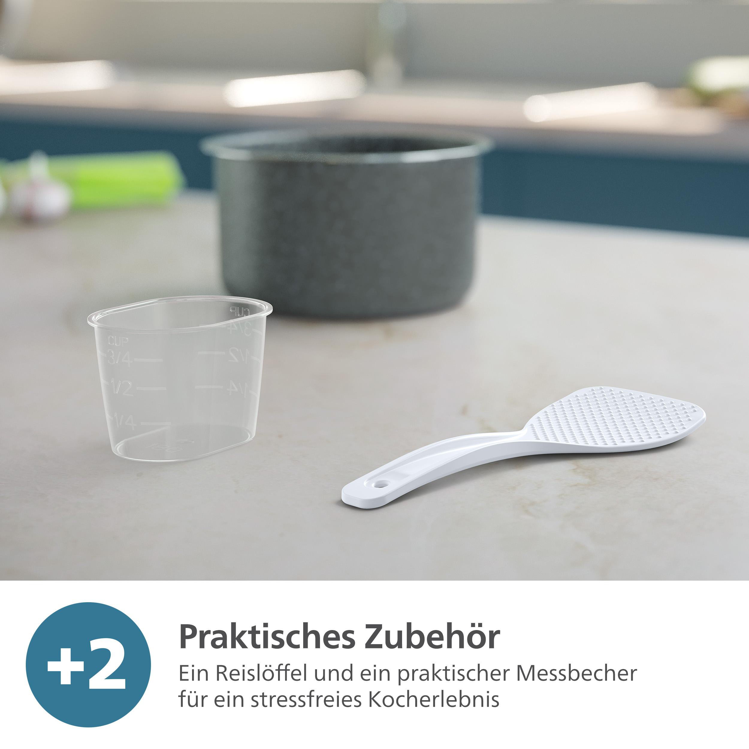Ein Messbecher und ein Reislöffel liegen auf einer Arbeitsplatte neben dem Reiskocher. Das Zubehör ist weiß.
