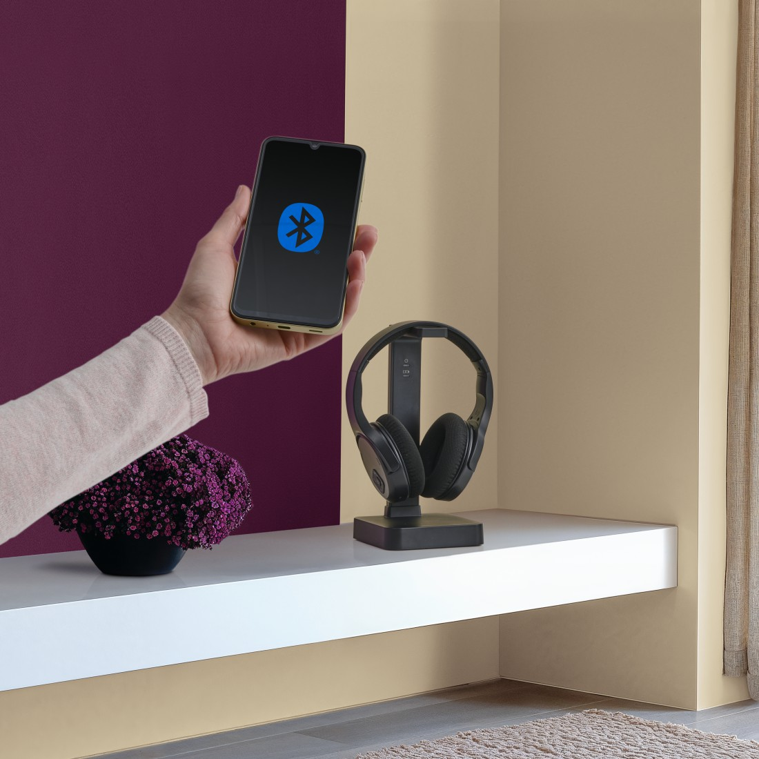 Eine Hand hält ein Telefon mit Bluetooth-Symbol. Kopfhörer liegen auf einem Ständer.
