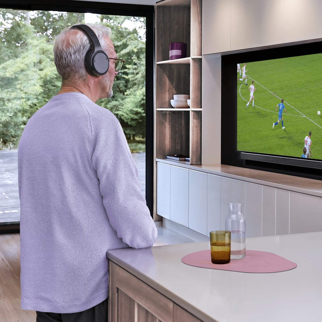 Un homme regarde la télé dans une cuisine. Il porte un casque et des lunettes. Foot à la télé.