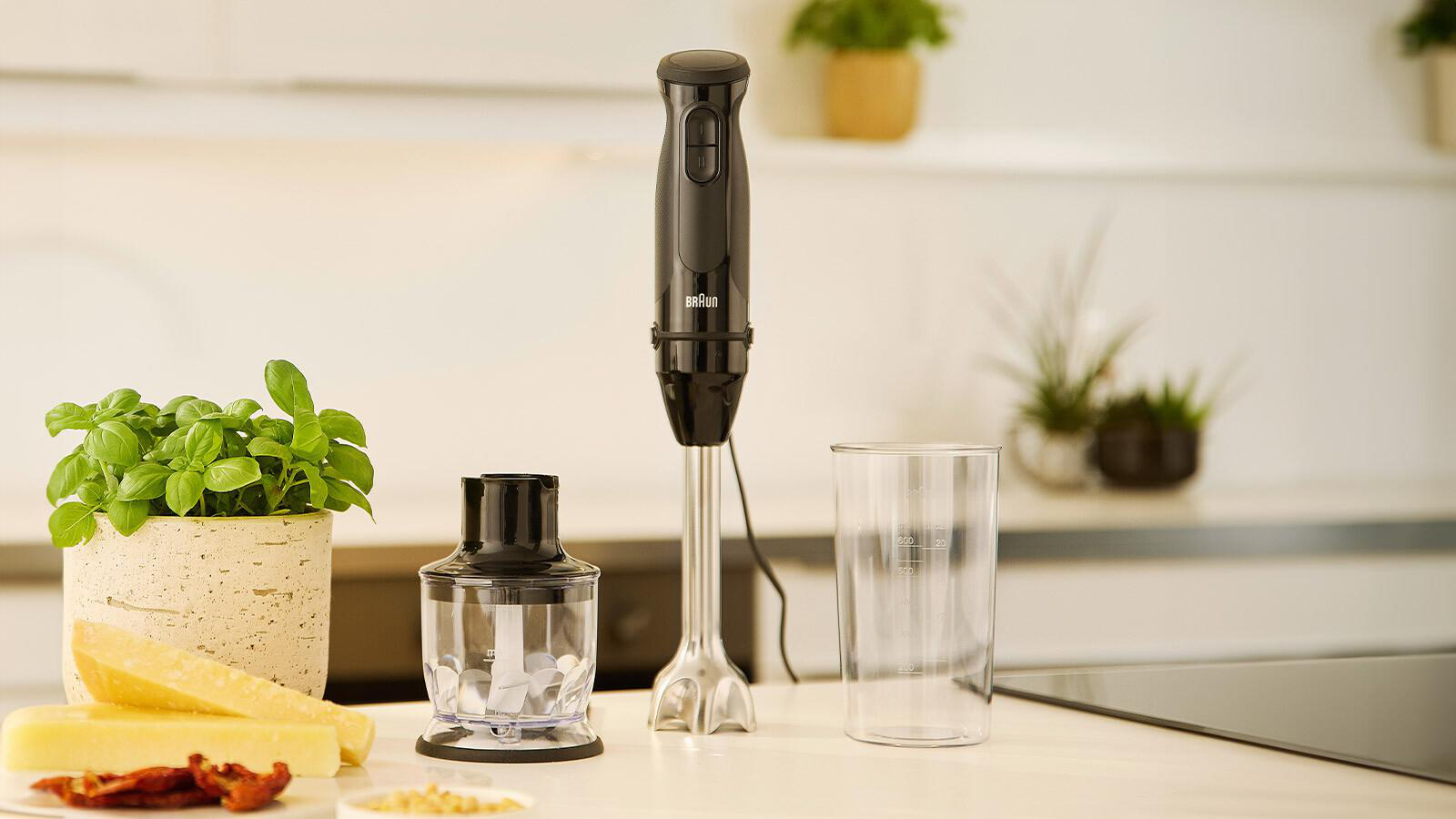Ein schwarzer Braun-Handmixer mit Zubehör, ein Glas, eine Basilikumpflanze und Käse auf einer weißen Oberfläche.
