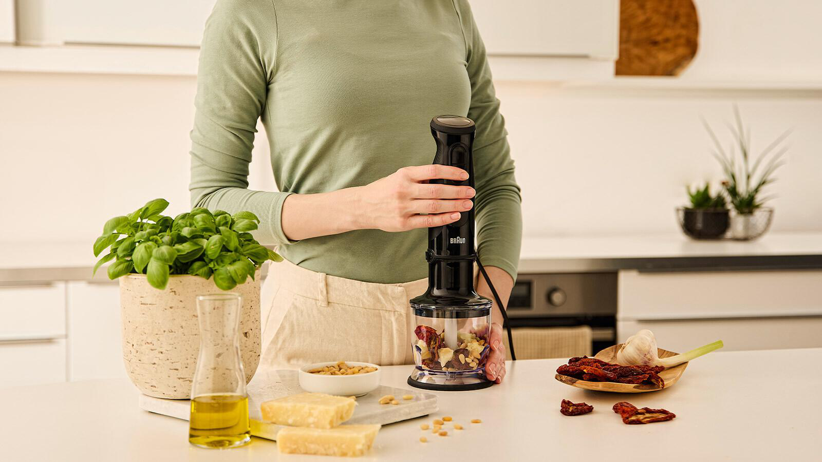 Eine Person hält einen schwarzen Stabmixer. Zutaten für Pesto auf der Theke: Basilikum, Öl, Pinienkerne, Käse und Tomaten.