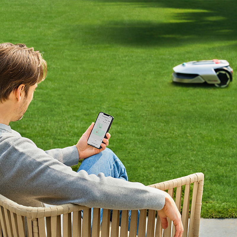 Oturan, telefon kullanan ve arka planda çimleri biçen bir robot çim biçme makinesine bakan adam.