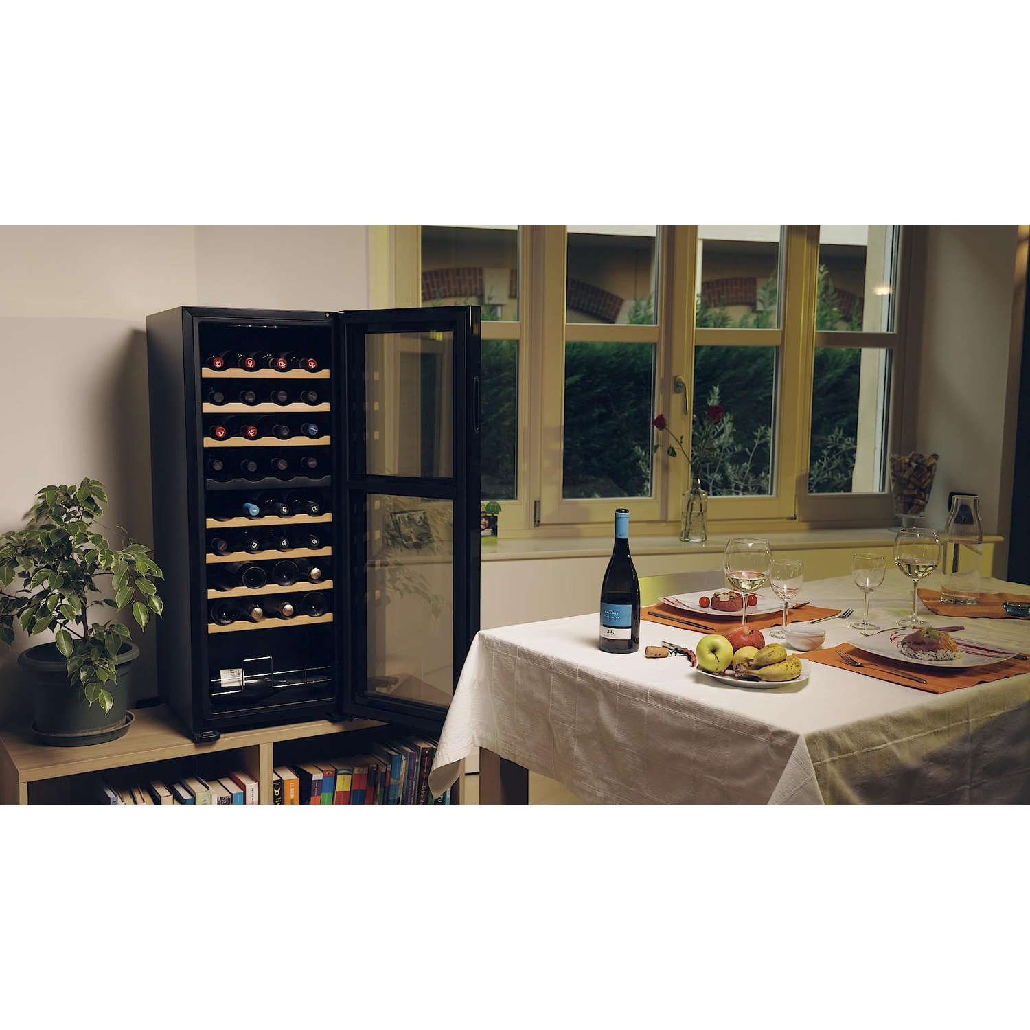 Una cantinetta con porta aperta, tavolo apparecchiato con vino, bicchieri, frutta e pianta.