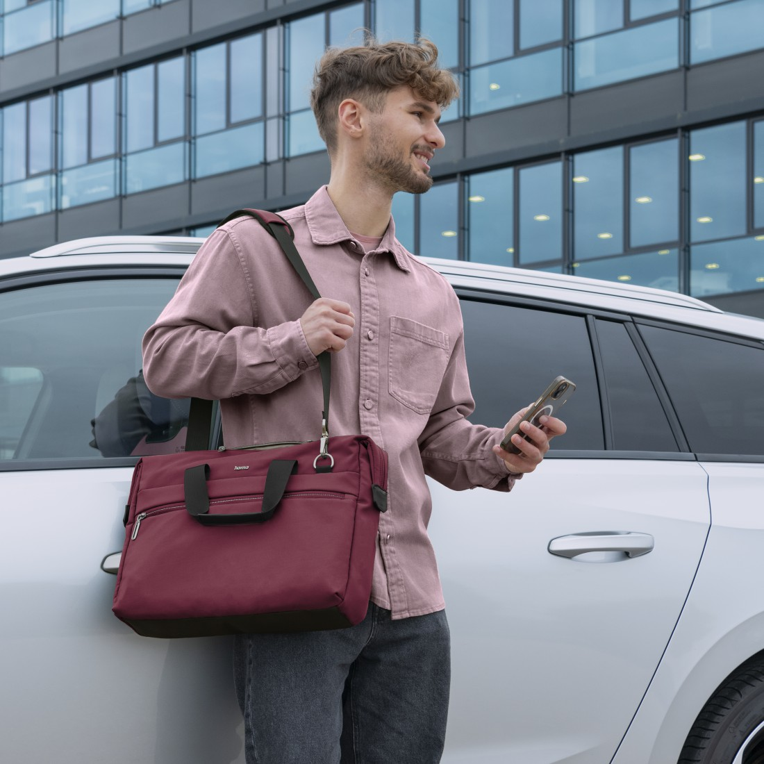 Mann mit weinroter Tasche geht in der Nähe eines weißen Autos und hält ein Telefon.