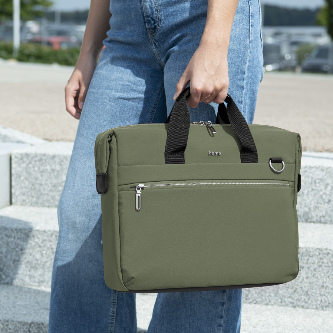 Eine Person hält eine grüne Laptoptasche. Sie trägt Jeans und steht vor Treppen.