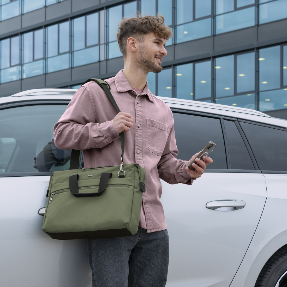 Mann im rosa Hemd, trägt olivgrüne Tasche, hält Telefon, neben weißem Auto und Glasgebäude.